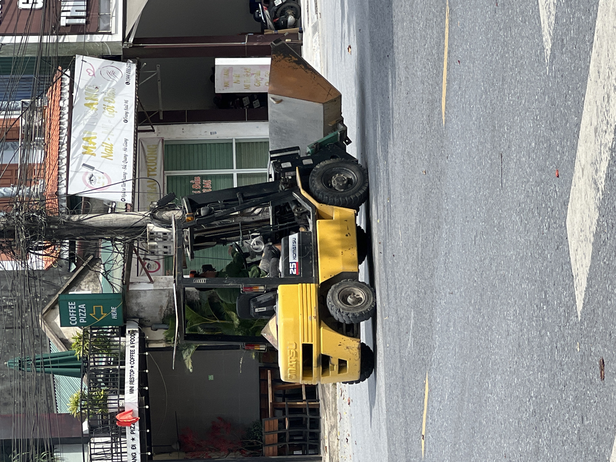 A yellow Komatsu forklift is parked on a paved road in what appears to be a commercial district in Tân Quang Commune, Vietnam. The forklift is positioned in the foreground, with its front facing left. A metal bin is attached to the lifting mechanism of the forklift, angled downwards. The forklift has a visible "KOMATSU" logo and the number "25" on its side. A conical Vietnamese hat (nón lá) is visible on top of the forklift. In the background, buildings with various signs line the street, including a green sign with white lettering that says "COFFEE PIZZA HERE" pointing left, and a large white banner with Vietnamese text and graphics, possibly advertising a nail salon. Power lines and electrical cables are strung across the top of the image. The road surface is asphalt, marked with yellow lines. Scattered on the road are fallen leaves. Other elements in the background include outdoor seating at what might be a cafe or restaurant, parked motorcycles, and architectural details of the buildings.