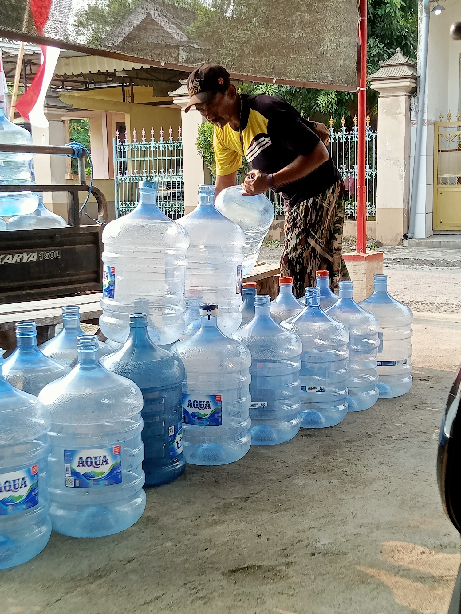 A man, wearing a dark baseball cap, a yellow t-shirt with black stripes on the sleeves, and patterned pants, is seen outdoors in the city of Simo, Indonesia. He is in the process of handling large, clear plastic water jugs. The jugs are mostly filled with clear liquid and are arranged on the ground in several rows. Some of the jugs have blue caps, while others have orange caps. One jug prominently displays the "AQUA" logo. The man appears to be transferring water or refilling a jug, as he is holding a smaller container or a cap over one of the large jugs. In the background, there is a structure that appears to be a building with a dark roof and a light-colored wall. There is also a metal fence with decorative elements and some greenery visible. A portion of a vehicle, possibly a car, is visible on the far right side of the image. The scene suggests a water refill or delivery point.