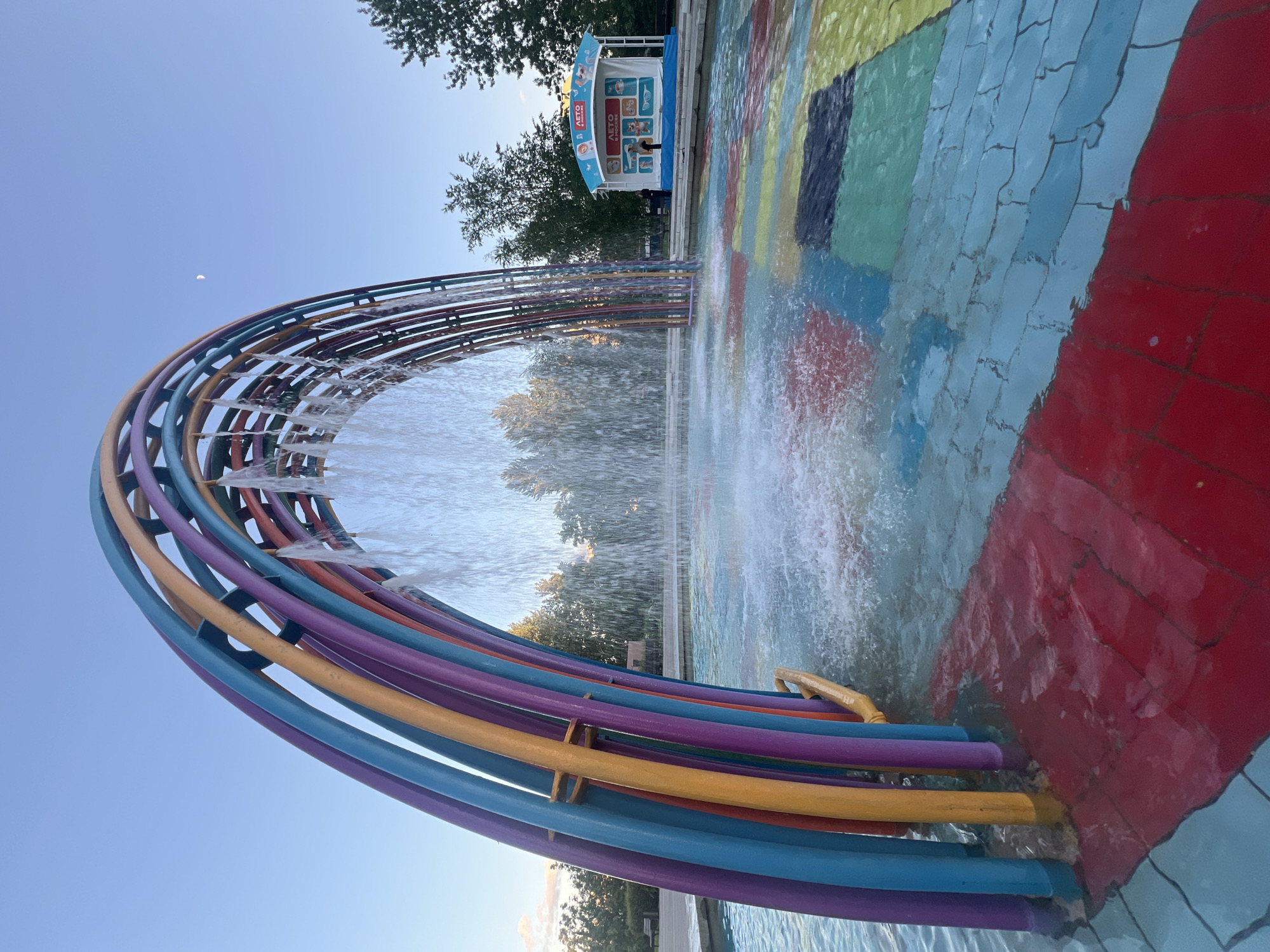 A rainbow-colored water fountain is flowing into a pool of water with colorful, tiled squares in Gorky Park, Moscow, Russia. The fountain consists of multiple curved metal pipes painted in different colors forming a rainbow shape. The water is spraying out of the pipes. The pool has a pattern of multi-colored tiles. In the background is a building with promotional material written in Russian. A crescent moon is visible in the sky.