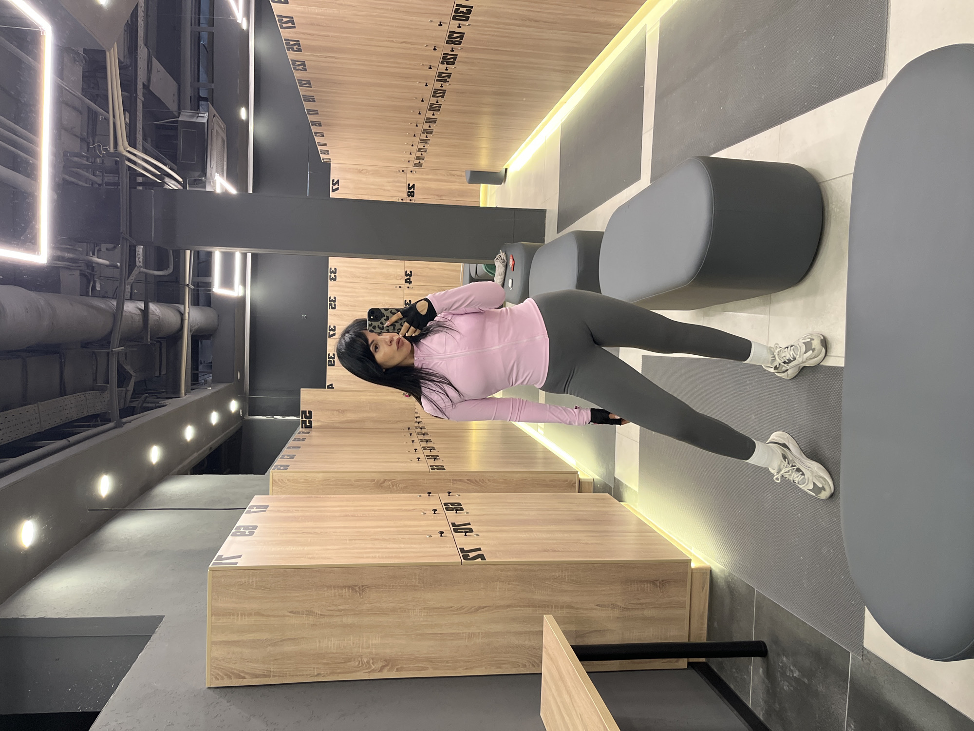A woman is positioned in a locker room interior, taking a selfie. She is wearing a pink long-sleeved shirt and gray leggings, along with white socks and athletic shoes. The locker room features rows of lockers with numbers on them. There are also gray, oval-shaped benches in the space. The lighting is a combination of natural and artificial, illuminating the scene. The location appears to be in Sochi, Russia.