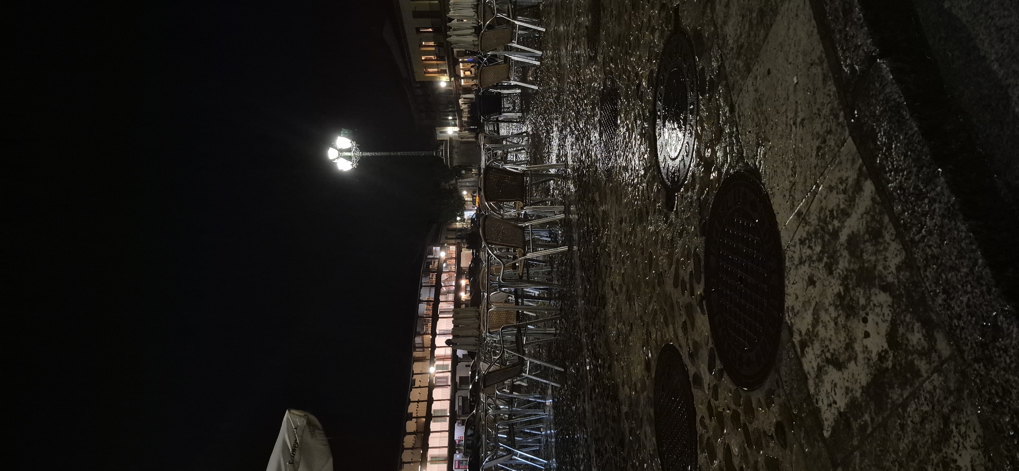 The image shows an outdoor cafe scene at night in the Plaza de Segovia, Navalcarnero, Spain. The cobblestone ground is wet, reflecting the lights from the surrounding buildings and a lamppost in the center of the plaza. Numerous tables and chairs, made of metal and wicker, are arranged in rows, many of them empty. In the background, a long, multi-story building with arcades and balconies is illuminated from within, suggesting shops or residences. A few indistinct figures can be seen through the windows of the building. The overall atmosphere is quiet and deserted, likely due to the late hour and wet conditions.
