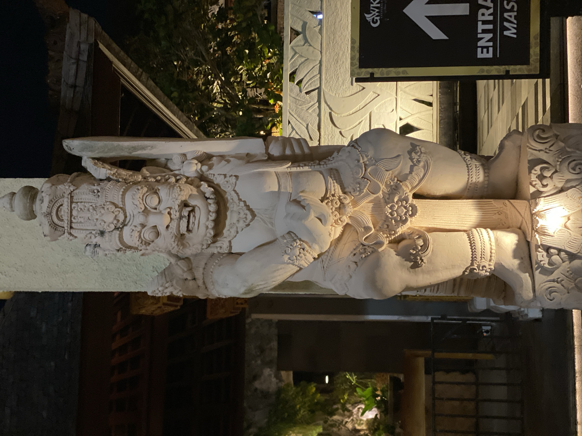 The image shows a nighttime view of a carved stone statue of a fierce, large figure standing in front of a textured wall and a sign. The statue is intricately detailed with a large headdress, a fierce facial expression with bared teeth, muscular arms, and ornate jewelry on its legs. It appears to be holding a sword or large weapon in its right hand. The statue is illuminated by artificial lights, casting shadows that highlight its texture.
To the right of the statue, a black sign with gold trim displays an upward-pointing arrow and text that reads "GWK CULTURAL PARK" and "ENTRANCE MASS..." The sign indicates a directional or informational purpose.
In the background, there is a glimpse of trees with illuminated foliage and architectural elements of a building, suggesting an outdoor setting at an establishment like a cultural park or resort. The overall impression is of a decorative or guardian statue at an entrance.
This statue is located at the Garuda Wisnu Kencana (GWK) Cultural Park in Ungasan, Kuta Selatan, Bali, Indonesia. GWK is a renowned landmark known for its massive statues and cultural performances. The statue depicted is likely a representation of a guardian deity or a character from Balinese mythology, often seen at the entrances of temples or significant cultural sites in Bali.