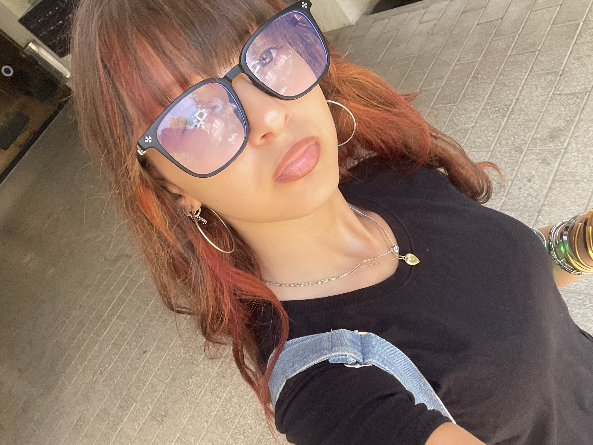A high-angle, close-up, selfie-style shot shows a person wearing sunglasses and a black t-shirt with denim suspenders. The person has reddish-brown hair with pink highlights and is wearing hoop earrings and a necklace with a heart-shaped pendant. The background is a brick-paved walkway, suggesting an outdoor urban setting in Raanana, Israel. The lighting is bright, casting reflections on the sunglasses, which show a blurry image of what appears to be a building or structure. The person's face is partially obscured by the sunglasses. Several bracelets are visible on the person's wrist.