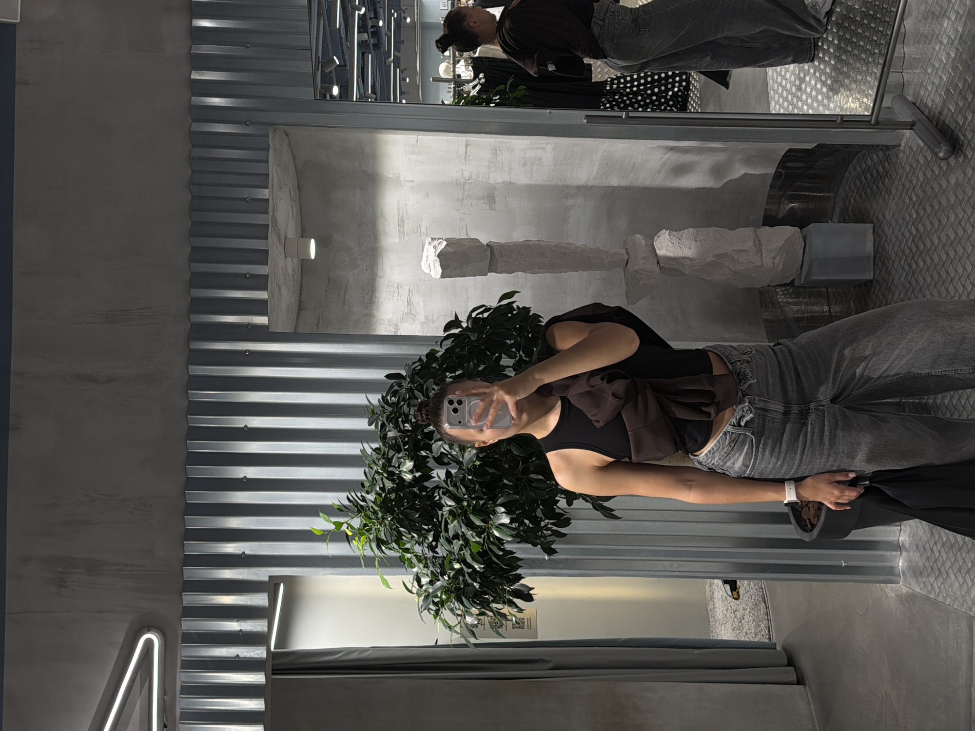 A young woman is taking a selfie in a store. She is wearing a black tank top, a brown sweater draped over her shoulders, and grey jeans. She has her hair in a bun and is holding a smartphone up to take the photo. She is standing in front of a large potted plant with dark green leaves.
The store has a modern, industrial aesthetic. The walls are a mix of corrugated metal and textured concrete. There are some artistic sculptures made of stone on display. In the background, there is a rack of clothing visible in a mirror. The lighting in the store is a mix of track lighting and neon accents. The floor is a textured, metallic material.
The image appears to be taken during the day, as indicated by the general lighting. The woman's expression is neutral, focused on taking the selfie. There is a QR code visible on a piece of paper near the plant, but the text is illegible. The location is identified as Moscow, Russia.
