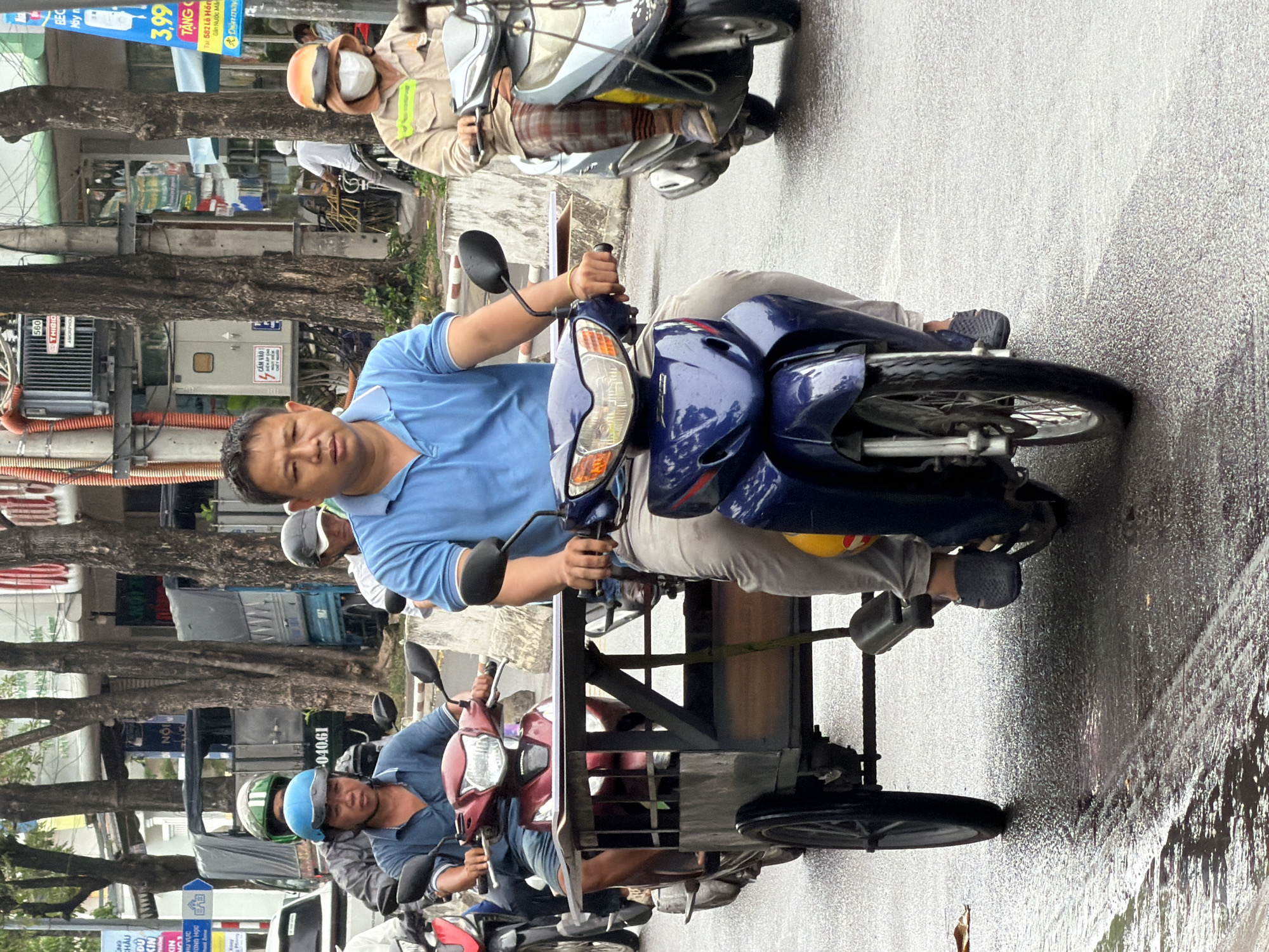 A medium shot captures a bustling street scene in Nha Trang, Vietnam, focusing on a man riding a blue motorcycle. He is wearing a light blue polo shirt and light-colored pants. The motorcycle has a metal cage attached to its front, indicating it may be used for transport or delivery. The road is wet and reflective, suggesting recent rain. Behind the main subject, other motorcyclists are visible, some wearing helmets and face masks, a common sight in urban Vietnam. Buildings line the street, some adorned with signage. The overall atmosphere is one of everyday urban life, with traffic and pedestrians moving along.