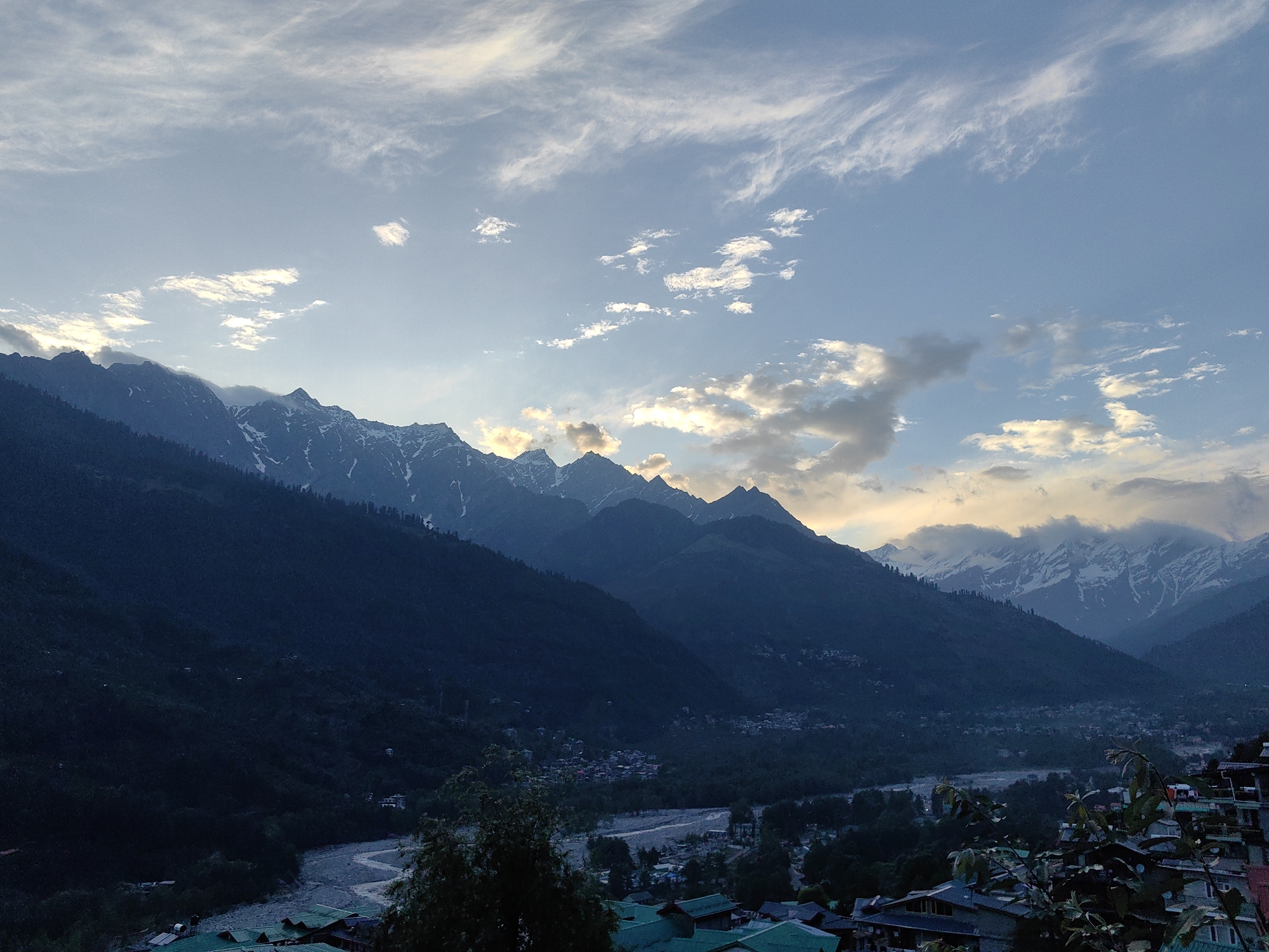 The image depicts a mountain landscape in Vashisht, India. The sky is partly cloudy with a mix of light blue and white clouds. Below, a series of dark, forested mountains stretches across the frame. Some peaks are capped with snow. In the valley, a river snakes through the landscape, with houses visible along its banks. In the foreground, rooftops of buildings are visible, indicating a settlement.