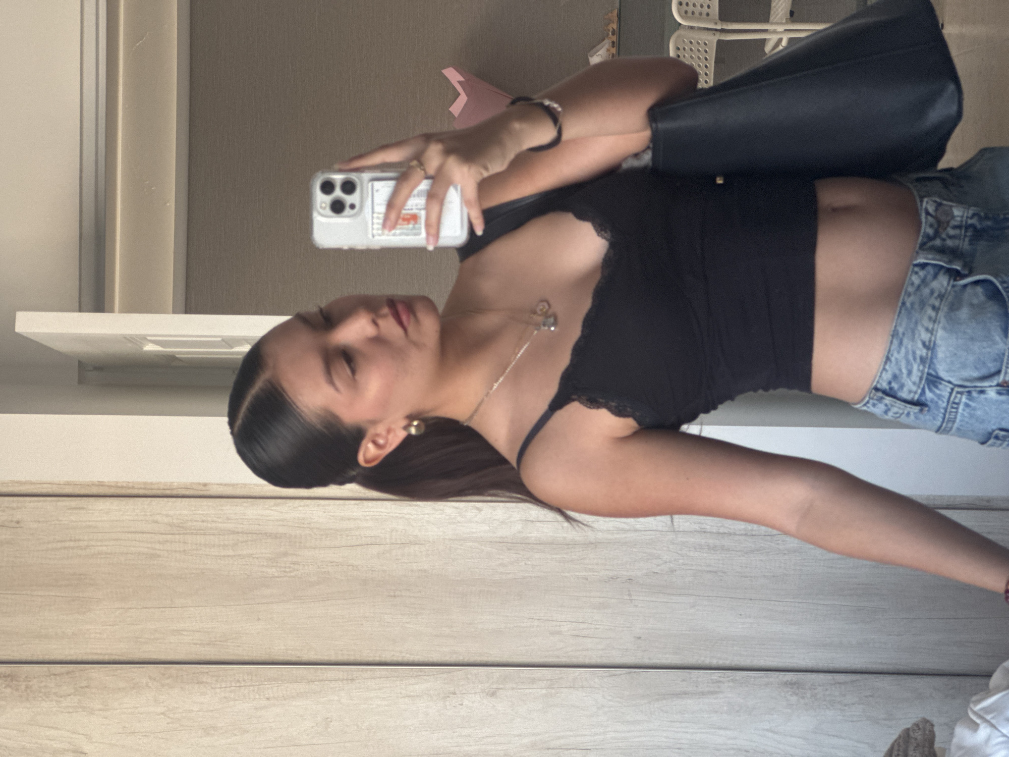 A young woman is taking a selfie in a mirrored location. She is holding a phone with a white case and multiple camera lenses. She is wearing a black cropped top with lace trim, blue denim jeans, and jewelry including a bracelet and a necklace. A black handbag is slung over her shoulder. The location appears to be indoors with light wooden paneling on the wall behind her. A white shelf and a beige wall are also visible. Based solely on the image, there is no indication as to whether this image was taken in San Francisco Ocotlán (Ocotlán), Mexico.