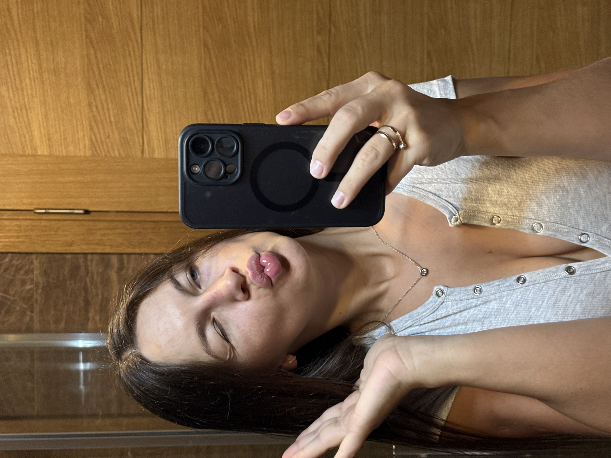 The image shows a woman taking a selfie in a mirror. She is wearing a grey, ribbed, button-up top that is unbuttoned low, revealing her cleavage. She has long, dark hair and is wearing a silver necklace and two rings on her left hand. She is winking and puckering her lips for the photo, and her left hand is raised with her palm facing the camera. She is holding a black smartphone in her right hand to take the picture. The background appears to be a bathroom with wooden walls and a reflective surface, possibly a shower door. The overall lighting is warm and natural. There is no indication of the location being Donji Pobori, Montenegro.