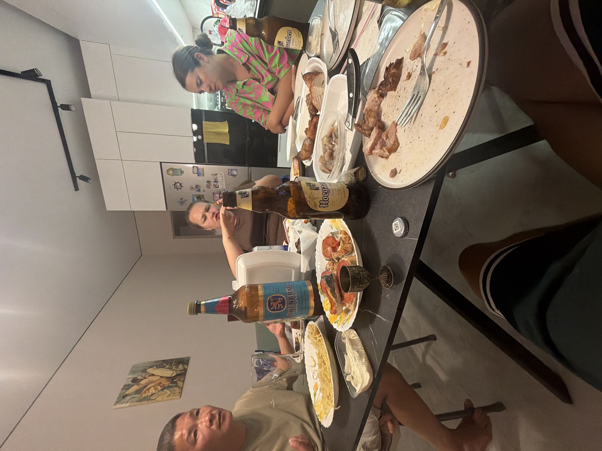 In an indoor setting, possibly a dining area or kitchen, four individuals are gathered around a dark, reflective table laden with food and beverages. The scene appears to be a meal in progress or recently concluded.
In the foreground, the legs and lower torso of a person wearing dark shorts with white stripes are visible. The table surface is cluttered with several plates of food, including what appears to be cooked meat, fish, and pasta or noodles. Empty and partially empty bottles of beer are prominently displayed. A glass and a small decorative goblet are also on the table. One bottle, a Löwenbräu, features a blue and gold label. Another, a Hoegaarden, has a white label with floral motifs. A white plastic container holds a creamy substance, possibly a dip or sauce. A bottle cap is visible on the table.
To the left, a man with short dark hair and a light-colored t-shirt is seated, his hands resting on the table. His gaze is directed forward, and his mouth is slightly open. Behind him, a partially visible glass can be seen.
In the center of the image, a woman with blonde hair pulled back is seated, her head tilted down as if looking at something on the table. She is wearing a brown top.
To the right, a woman with dark hair tied up is seated, wearing a vibrant green and pink patterned shirt. Her arms are crossed, and she is looking to her right.
In the background, a modern kitchen area is visible, featuring white cabinets and appliances, including a refrigerator adorned with magnets and photographs. A track lighting system is mounted on the ceiling. A piece of artwork, depicting what looks like bananas or similar leaves in muted colors, hangs on the wall to the left. The overall lighting is artificial, contributing to a slightly dim but functional ambiance. The floor appears to be a light-colored material.