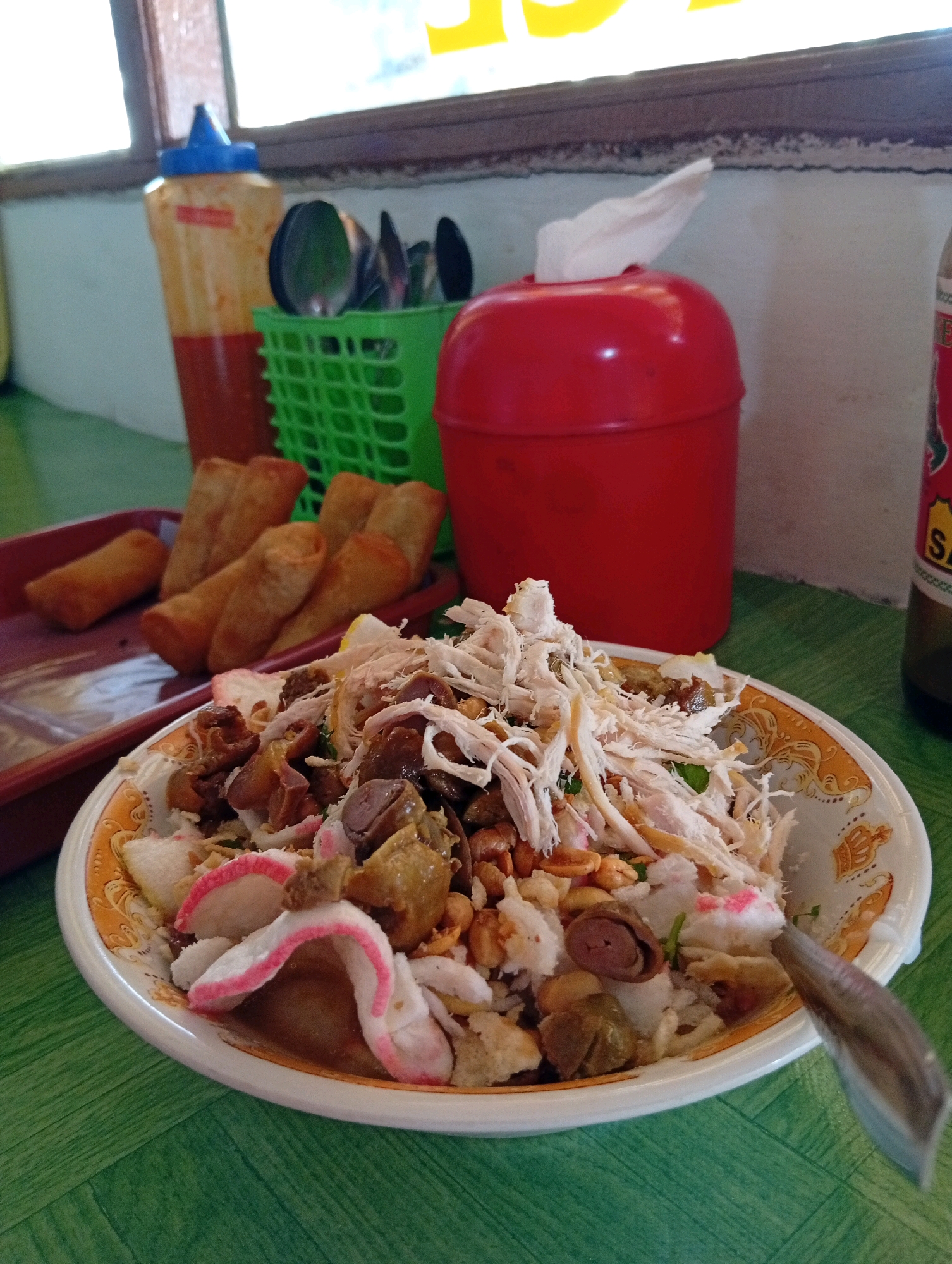 This is a close-up, eye-level shot of a bowl of food, likely chicken soup or a similar dish, placed on a green textured table. The bowl is white with an orange and yellow decorative rim. The dish contains shredded chicken, rice, some dark pieces that appear to be chicken innards, and pink and white puffed crackers. A spoon with a metallic finish is partially submerged in the food on the right side.
In the background, a red plastic container with a white tissue protruding from the top is visible. To the left of that, a green plastic basket holds several spoons and knives. Behind the basket, there is a plastic squeeze bottle with a red and orange design, likely a condiment bottle. Further to the left, part of a window frame and a window are visible. On the far right, the neck and label of a dark glass bottle, possibly beer or a soft drink, are partially visible. The overall setting appears to be a casual dining environment. The location is identified as Cipendawa, Indonesia.