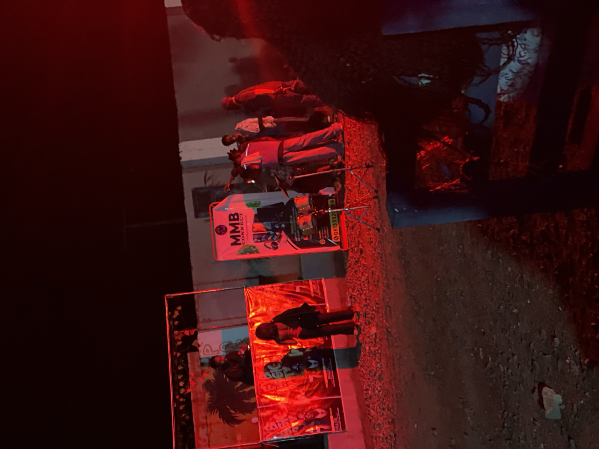 This image depicts an outdoor event at night in Durumi, Nigeria. The scene is illuminated by red lighting, casting a vibrant glow on the surroundings. In the background, a raised stage or platform is visible, with individuals appearing to be performing or operating equipment. Banners with text and graphics are displayed, one of which prominently features "MMB CONNECT". There are musical instruments, including drums, set up on the ground. Several people are present, some standing on the platform and others on the ground. The ground is covered in gravel and dirt. In the foreground, a person is partially visible, seated and facing away from the camera. The overall atmosphere suggests a lively gathering or performance.