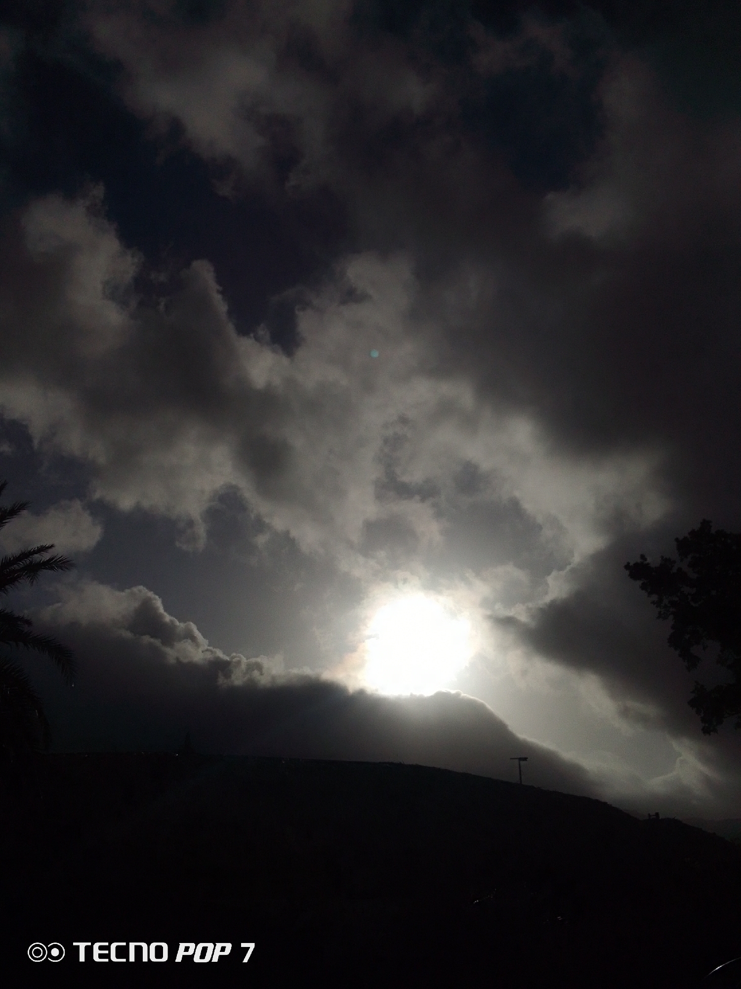 The image captures a dramatic sky in Pedro González, Venezuela. Overcast clouds obscure much of the sky, with the sun attempting to break through, creating a bright glow that permeates the cloud cover. In the foreground, a dark, silhouetted landscape, possibly a hill or elevated terrain, is visible. A faint streetlight pole is discernable on the ridge. The image is taken from ground level, looking upwards. The bottom left corner contains the watermark "TECNO POP 7". A palm tree is visible on the left side. The presence of trees on the right side suggests vegetation typical of the location.