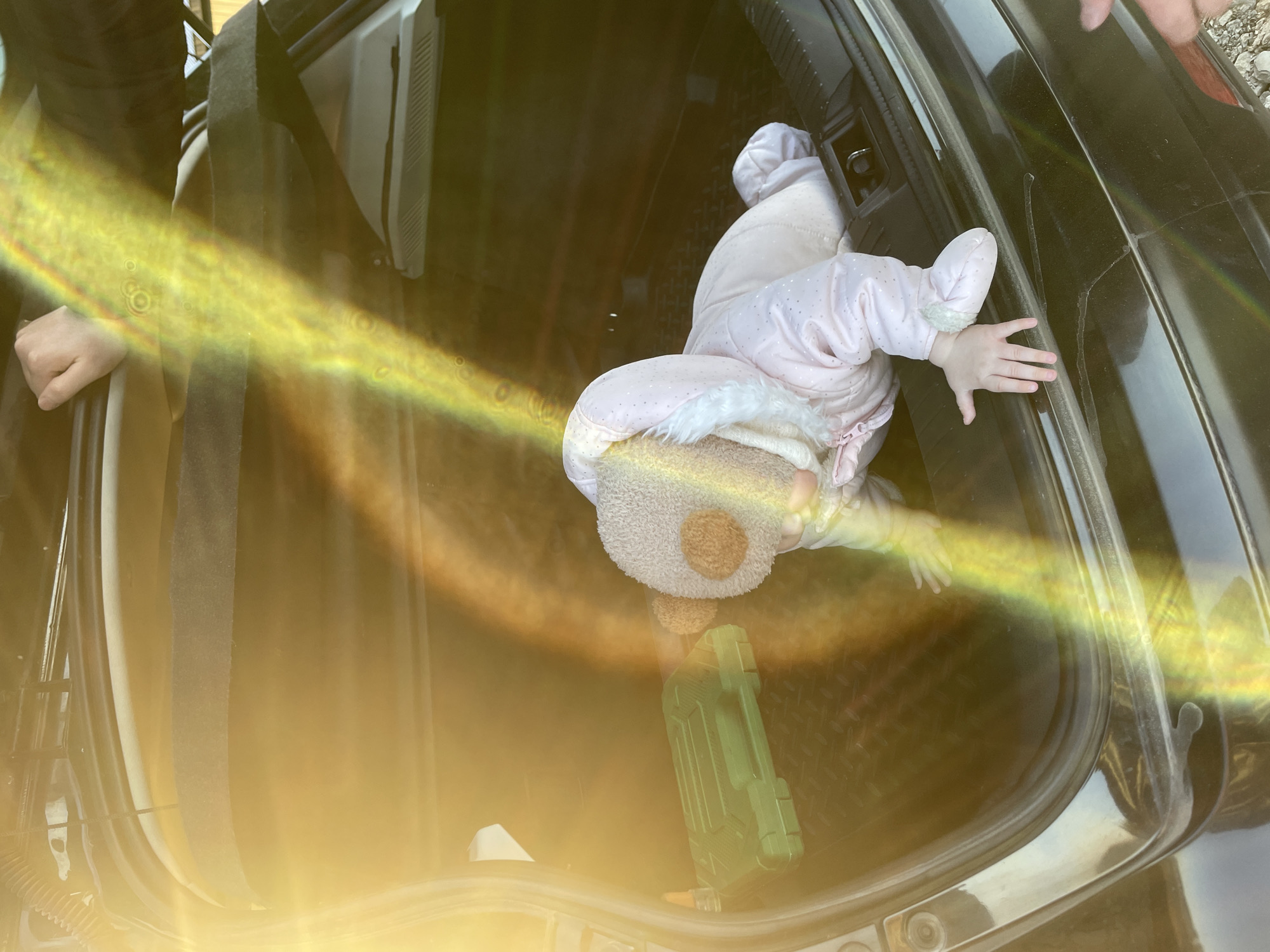 The image depicts a baby or toddler playfully interacting within the open trunk of a black car. The child is dressed in a light pink padded suit adorned with white fur trim and a charming teddy bear-shaped hat. The baby is positioned face-down, seemingly exploring the trunk's interior and reaching out towards the back of the vehicle with both hands.
The car's trunk features a dark interior with a textured mat. A distinctive green, rectangular plastic toy, resembling a tool case, is visible near the child's head at the bottom of the trunk. On the left side of the frame, an adult hand is partially visible, possibly holding a strap or leaning on the car's interior. Another adult hand can be seen in the upper right, resting on the car's exterior, which appears black and has some visible streaks or dust. The ground outside the car in the top right corner consists of gravel or dirt.
The scene is illuminated by strong natural sunlight, creating a noticeable yellowish-orange lens flare that spans diagonally across the middle of the image, adding a warm glow but also obscuring some details. The baby's warm attire suggests a cool outdoor temperature. The overall impression is one of a casual, playful moment. The setting is identified as Podshivalovo, Russia. No visible text is present in the image.