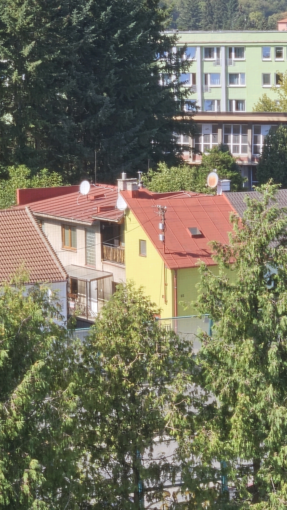 The image captures a residential area in Prešov, Slovakia, characterized by a cluster of houses and a larger apartment building. In the foreground, several trees with dense green foliage are visible, partially obscuring the view of the buildings. The houses have red tiled roofs, and one of them is painted yellow. One of the houses has a sunroom with a glass enclosure. Antennas and satellite dishes are mounted on the roofs of the houses, indicating typical residential installations. The apartment building in the background is painted light green with white window frames and balconies. The overall scene suggests a suburban or urban residential setting bathed in natural sunlight.