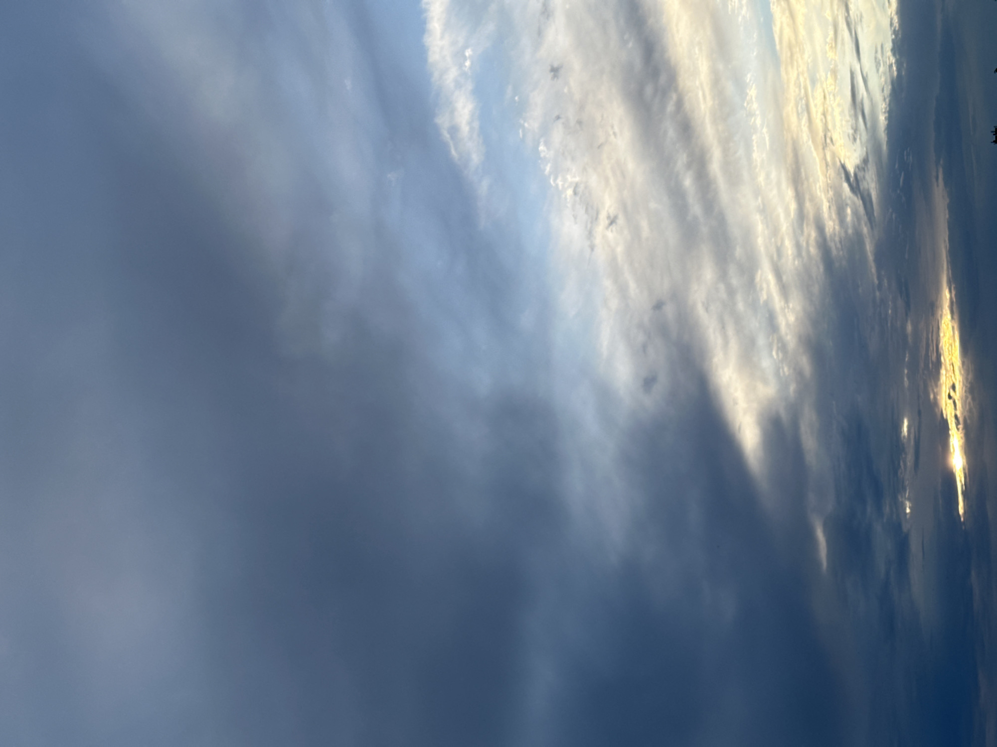 A vertically oriented photograph shows a sky with varied cloud formations. The left side of the frame is dominated by dark, thick clouds. Toward the right, the clouds are lighter, some appearing almost white and streaked. Bright, golden light shines through some of the clouds on the right side. At the far right edge of the image, there is a dark shape which may be trees or other vegetation. The location is Unknown, Unknown.