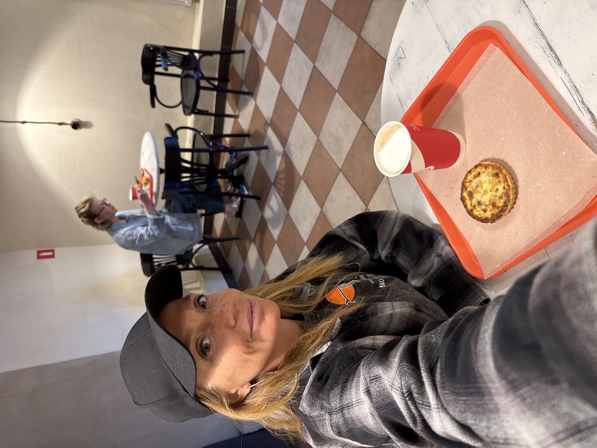 A woman is taking a selfie in a cafe with a checkered floor. She is wearing a black baseball cap and a plaid shirt. Her hair is blonde and she has green eyes. In the background, another woman in a light blue shirt is sitting at a table. There are black chairs and tables in the cafe. On a table in the foreground, there is a red tray with a pastry and a red and white cup of coffee. The image appears to be taken during the day. There is text on the woman's shirt that is partially visible. The context provided indicates the location is Vsevolozhsk, Russia.