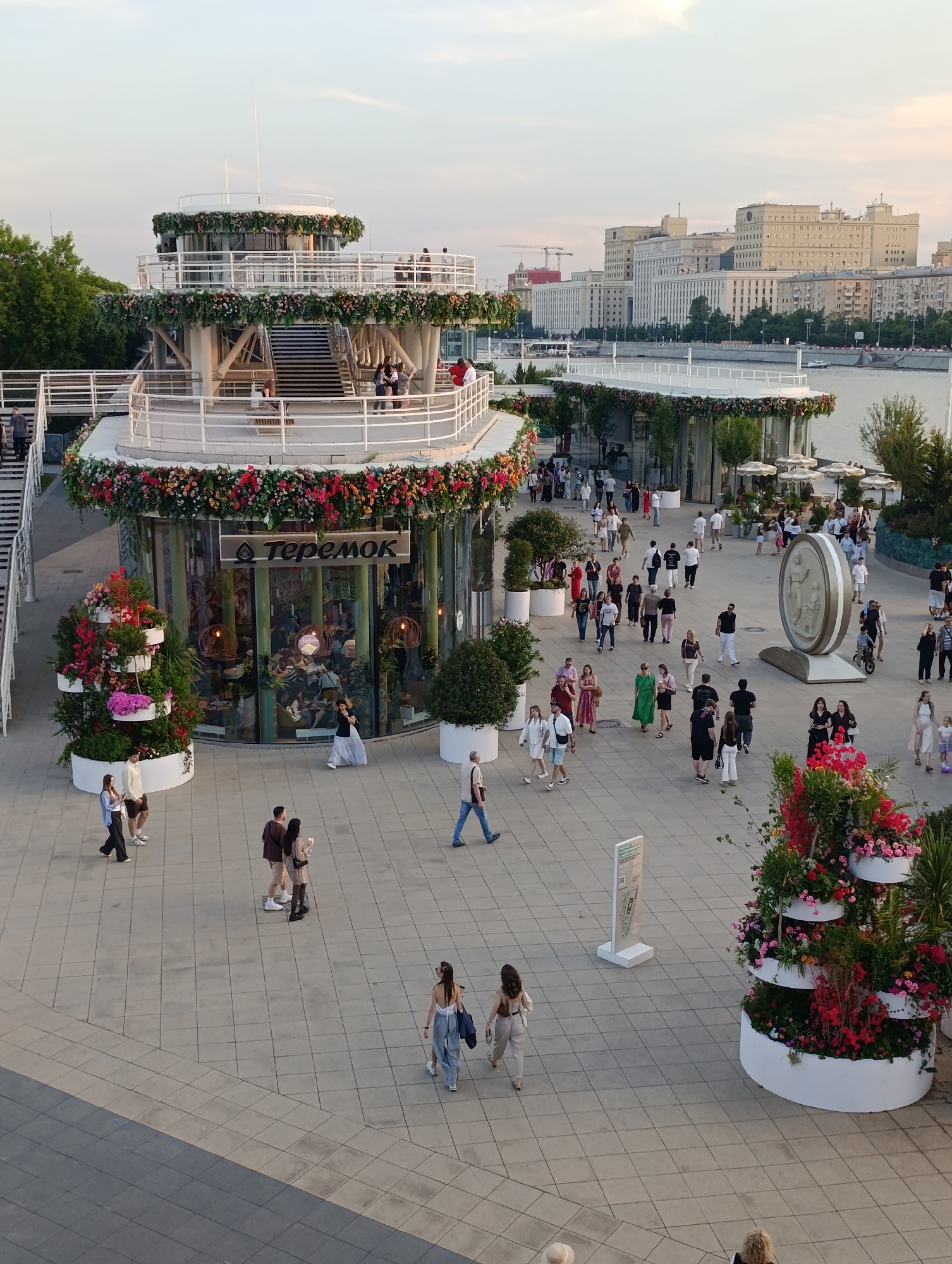 This image presents an overhead view of a bustling pedestrian area on Pushkinskaya Embankment, located within Gorky Park in Moscow, Russia. The Moscow River and the main building of the Ministry of Defence are visible in the background.
In the mid-ground, a multi-tiered, circular, white pavilion structure is prominently featured, extensively decorated with garlands of red, pink, and green flowers and foliage along its railings and edges. The ground level of this pavilion houses a "Теремок" (Teremok) restaurant, identifiable by its Cyrillic sign, with large glass windows revealing patrons seated inside. Open-air balconies on the upper tiers are also adorned with flowers, and several individuals are visible on them. White staircases connect the different levels of the pavilion.
The foreground and mid-ground depict a wide plaza paved with light gray square tiles, traversed by dozens of people. Individuals are observed walking, standing, and conversing in diverse groups and as solo figures, dressed in casual attire. Decorative elements include several large, white, tiered planters overflowing with vibrant red, pink, and purple flowers, and smaller green shrubs in white pots. On the right side of the plaza, a large, circular, pale gray sculptural monument with bas-relief figures stands on a plinth. Further back on the right, an outdoor seating area with white umbrellas and tables is visible. A white information stand is positioned in the lower-left area of the plaza.
The Moscow River is visible to the right in the background, with a few small boats on its calm surface. Across the river, a long, light-colored complex of multi-story buildings, characterized by numerous windows and a distinct architectural style, is identifiable as the main building of the Ministry of Defence of the Russian Federation on Frunzenskaya Embankment. The sky above is clear and light, indicating late afternoon or early evening, casting soft, elongated shadows. Green trees line the left periphery of the scene and the distant riverbank.