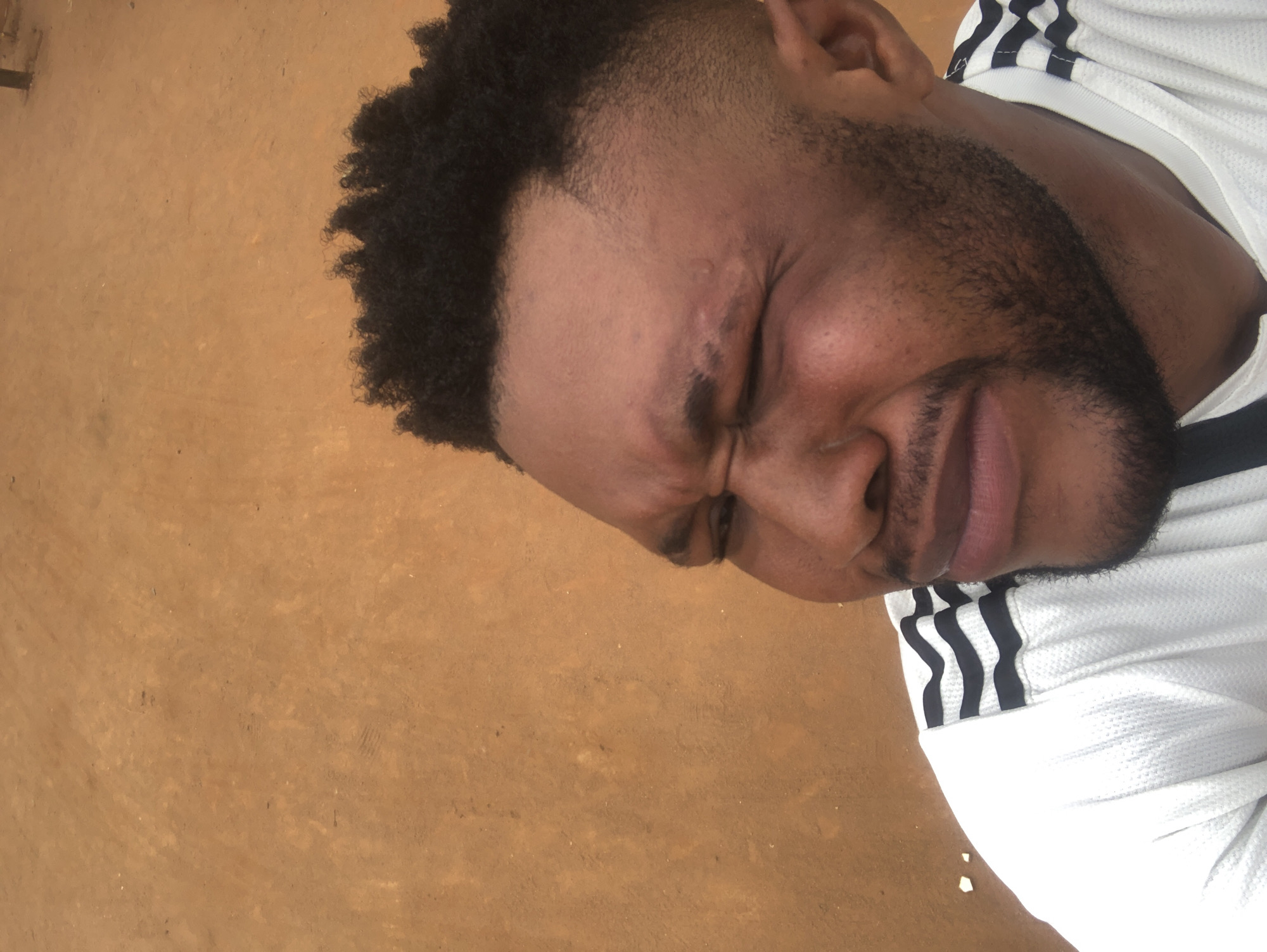 A close-up shot depicts a man with dark hair and a beard, wearing a white shirt with black stripes, lying on a surface of tan color. His face is contorted in a grimace or expression of discomfort, with eyes closed. The location appears to be an outdoor setting, but the specific location in the city of Unknown, Unknown cannot be determined from the provided image.