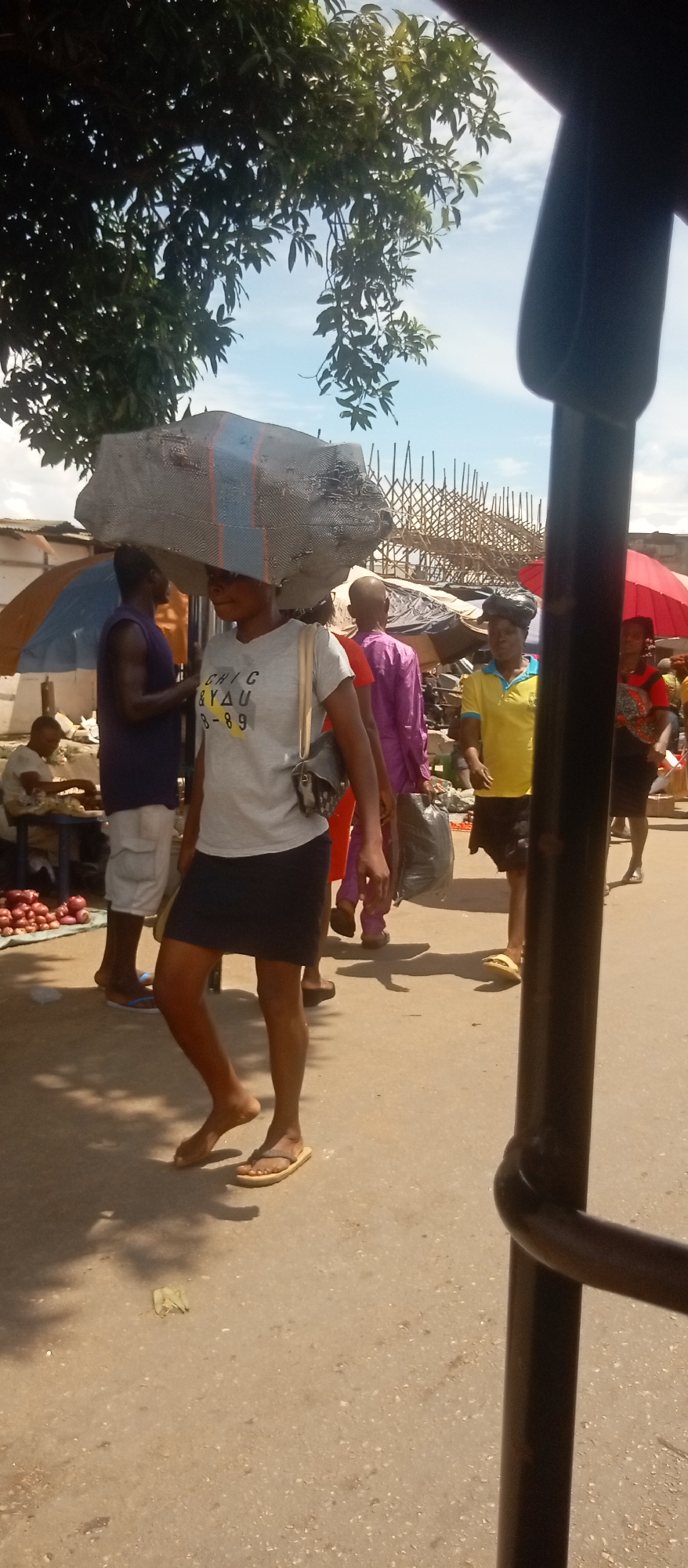 The image depicts a street scene in an outdoor market. A woman is carrying a large gray sack on her head while walking forward. She is wearing a gray t-shirt, a dark skirt, and sandals. She has a small purse over her shoulder. Other people are also walking along the street, including a man in a purple shirt and a woman in a yellow shirt. Vendors can be seen in the background selling their wares. The scene is bathed in sunlight with shadows cast on the ground. A black metal bar partially obscures the right side of the frame, suggesting the view is taken from inside a vehicle. Vegetation and market structures can be seen overhead.