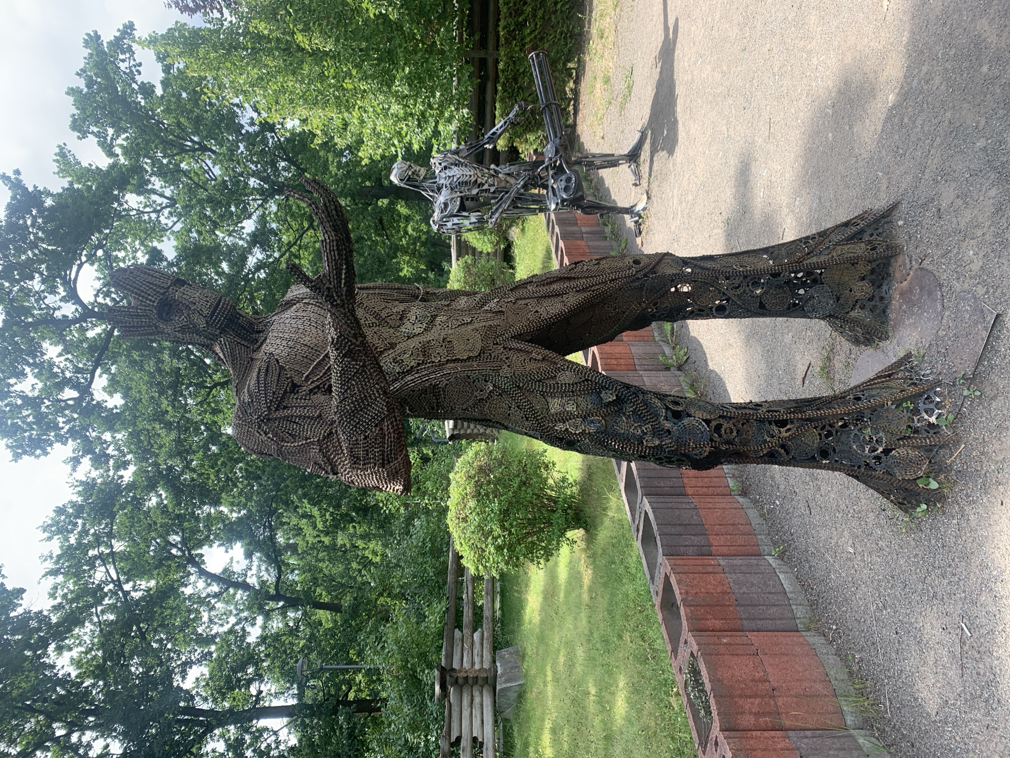 A large, humanoid sculpture made of metal gears and chains, resembling Groot from Guardians of the Galaxy, stands in the foreground. The sculpture is dark brown and has intricate details. To the right and slightly behind the Groot-like figure, a second sculpture of a skeletal robotic figure, reminiscent of the Terminator, is visible. This figure is made of polished metal and holds a large Gatling gun. The sculptures are situated outdoors in a park-like setting. Behind the sculptures, lush green trees and foliage form a dense backdrop. A wooden fence and a low, decorative wall made of reddish-brown and gray concrete blocks line the area around the sculptures. The ground is covered with gravel and dirt. The image appears to have been taken in Bucha, Ukraine, as evidenced by the location tag.