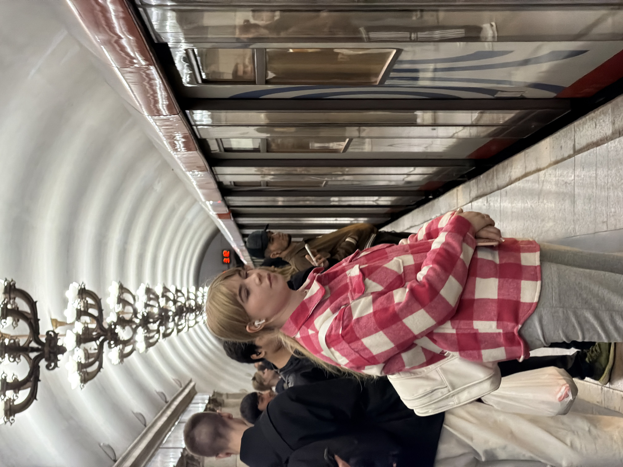 The image shows a group of people waiting on a platform in a Moscow Metro station. The station is identifiable by its vaulted ceiling and ornate chandeliers. A train is at the platform. A young woman in a red and white checkered shirt is standing in the foreground. Several other people are standing behind her. A man wearing a cap is smoking a cigarette.