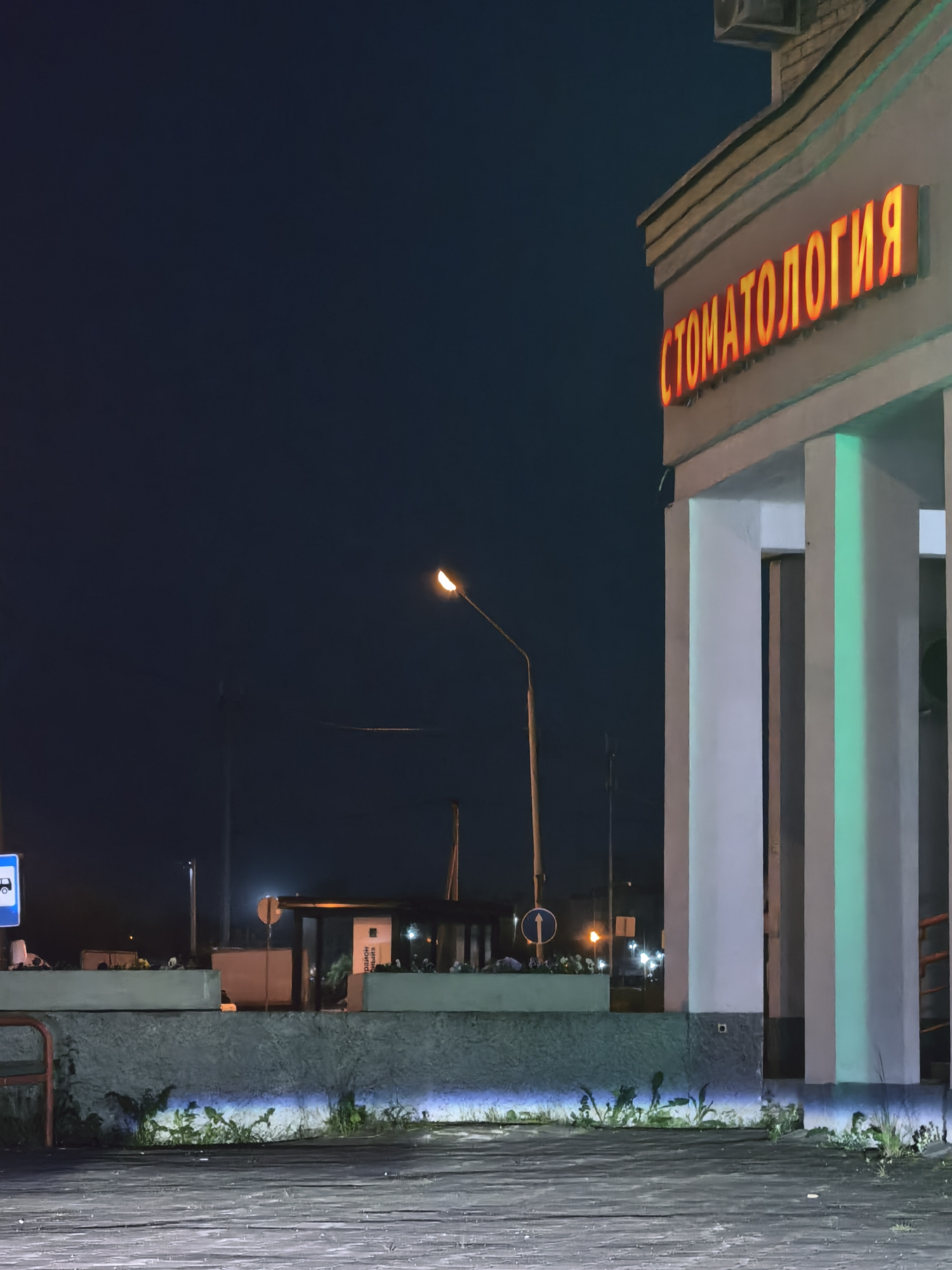 An exterior shot of a building at night shows a lit-up sign that reads "СТОМАТОЛОГИЯ" in large orange letters. The building has large white pillars that are lit with a green light. In the background, there are streetlights, a bus stop, and some greenery. The ground in front of the building is paved with a textured material.