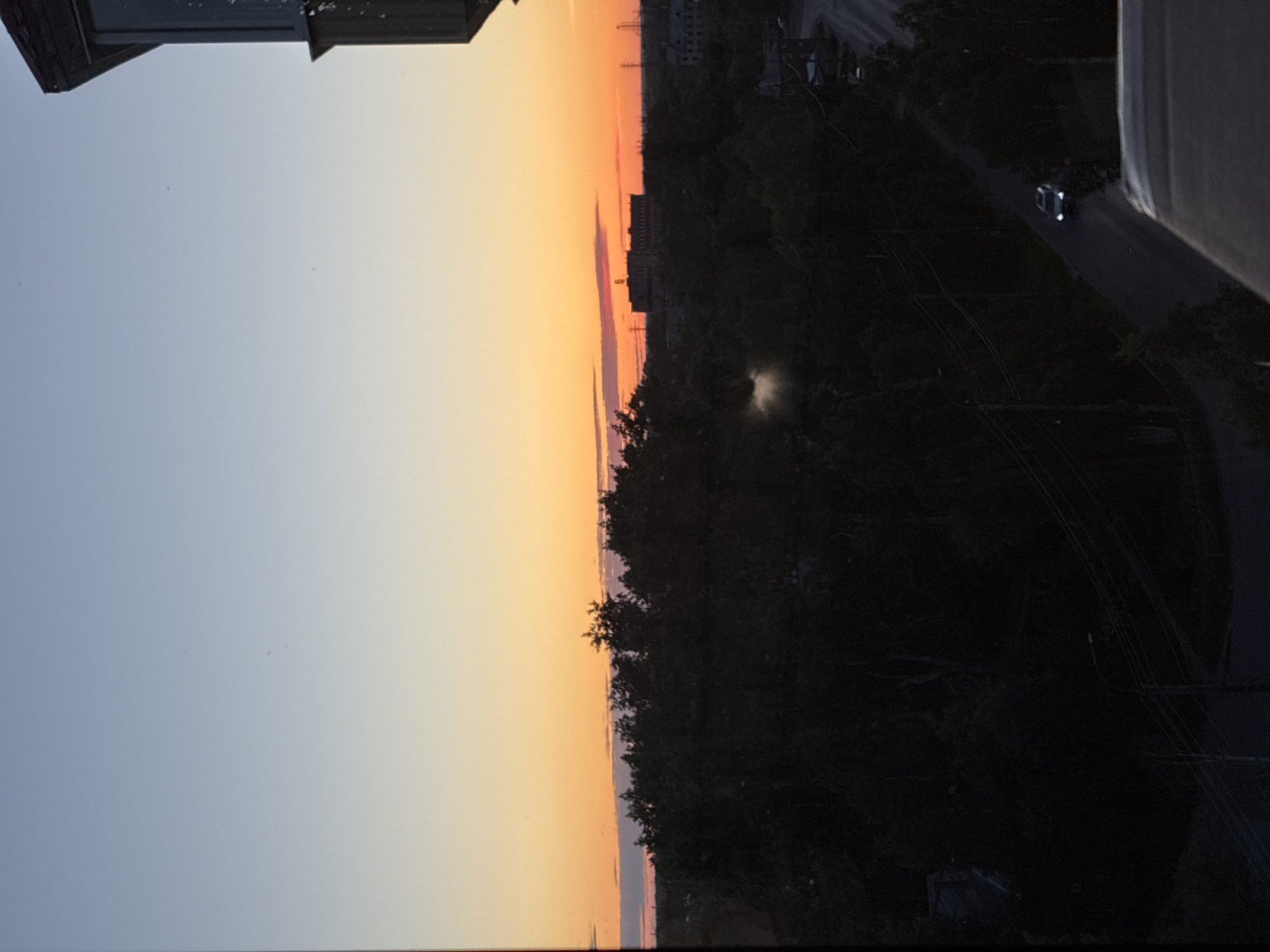 The image depicts a city view at dusk. The sky transitions from a pale blue at the top to a vibrant orange and pink near the horizon, suggesting a sunset or sunrise. Silhouetted against the colorful sky are dense trees that dominate the midground. In the distance, buildings and structures are visible, with one larger building appearing to be a factory or industrial complex. A road winds through the scene in the lower right, with a single white car driving on it. The overall impression is one of a somewhat urbanized landscape with significant natural elements.