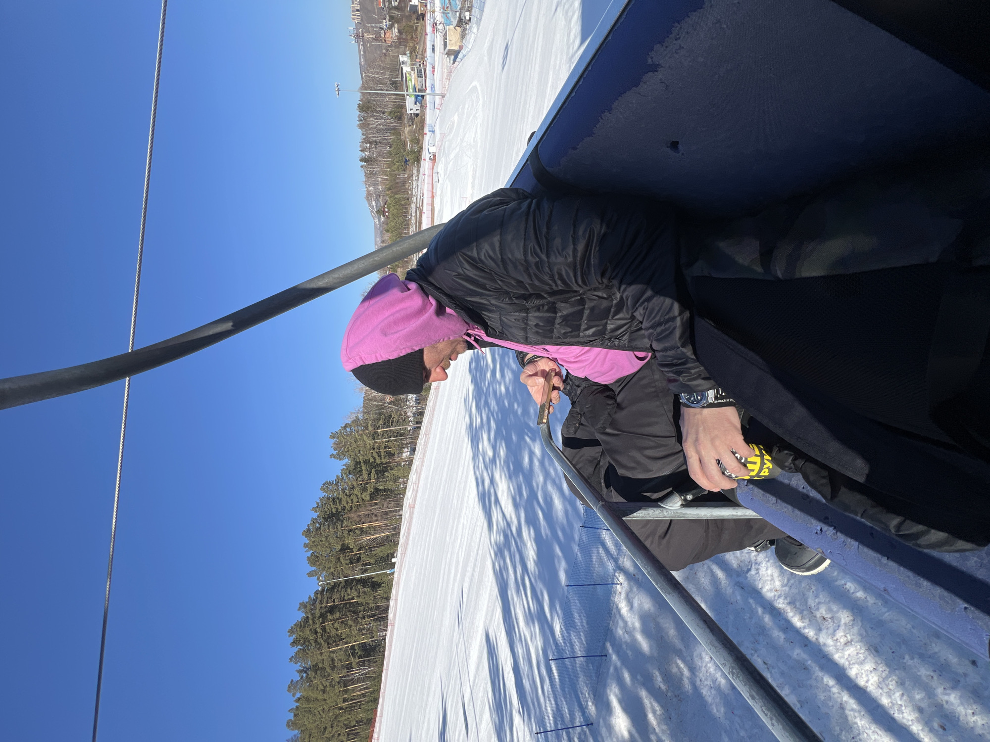 The image shows a person wearing a black jacket and a pink hood on a ski lift. They are holding a yellow and black object in their hands. The background consists of a snowy slope with trees, and a blue sky. The location is a ski resort, in the city of Unknown, Unknown.