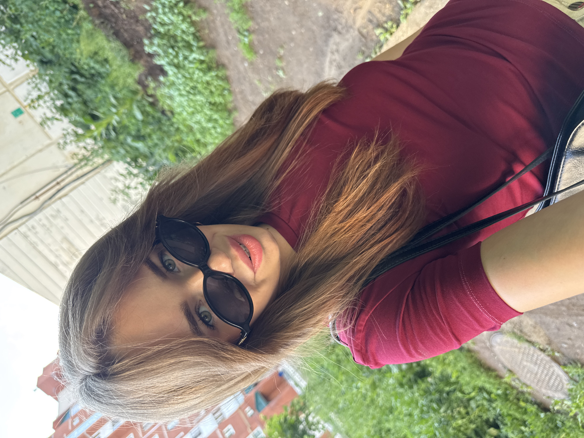 A young woman with long, light brown hair, wearing a dark red t-shirt and black sunglasses, takes a selfie outdoors. She is looking directly at the camera with a slight smile, her lips parted. The background shows a garden area with green foliage and a dirt path, suggesting an urban park or residential courtyard. In the upper right corner, a portion of a red brick building is visible. The image appears to have been taken during daylight hours. The scene is consistent with a casual outdoor setting in Chaikovsky, Russia.