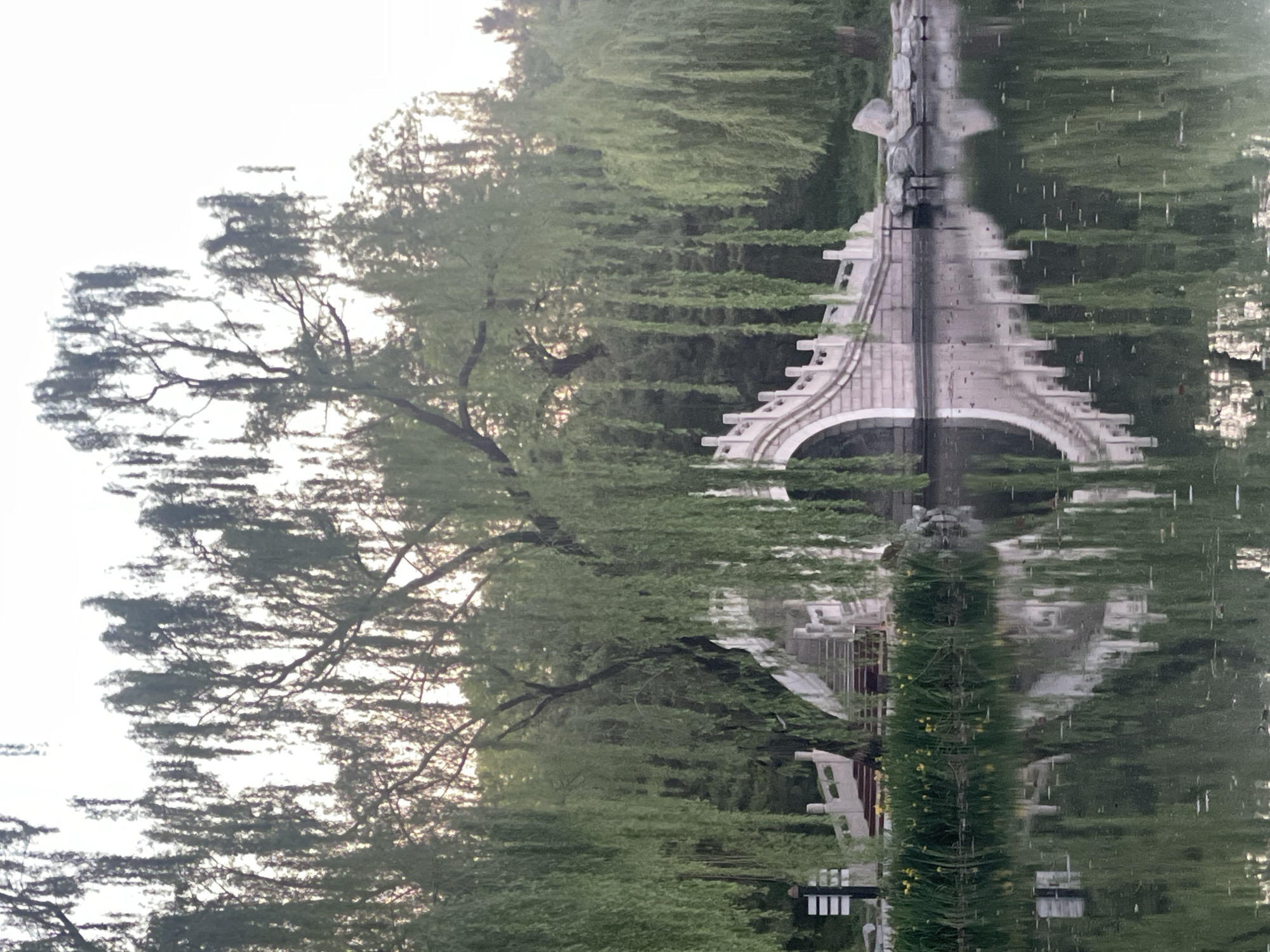 The image shows a reflection in water of a scene in Haidian District, China. The reflection features a stone bridge or structure with an arch, surrounded by trees with green foliage. There are street lights and buildings visible in the reflection. The sky above is light with faint clouds. The image is rotated.