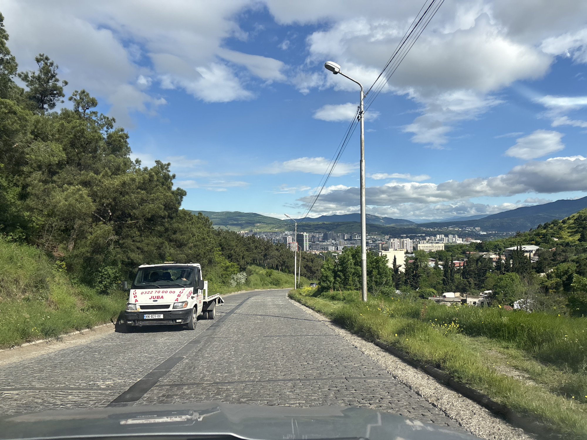 The image captures a view from within a vehicle driving on a cobblestone road in Tbilisi, Georgia. On the left side of the road, dense green trees and vegetation slope downhill. A white tow truck with "JUBA" printed in red block letters is parked facing the camera. The truck's license plate reads "KK-029-RR." The road curves slightly uphill to the right, flanked by a grassy verge. In the distance, a cityscape with numerous tall buildings is visible against a backdrop of mountains and a blue sky with scattered clouds. A tall street lamp stands along the road. The foreground shows a partial view of the vehicle's dashboard or hood from which the image is taken.