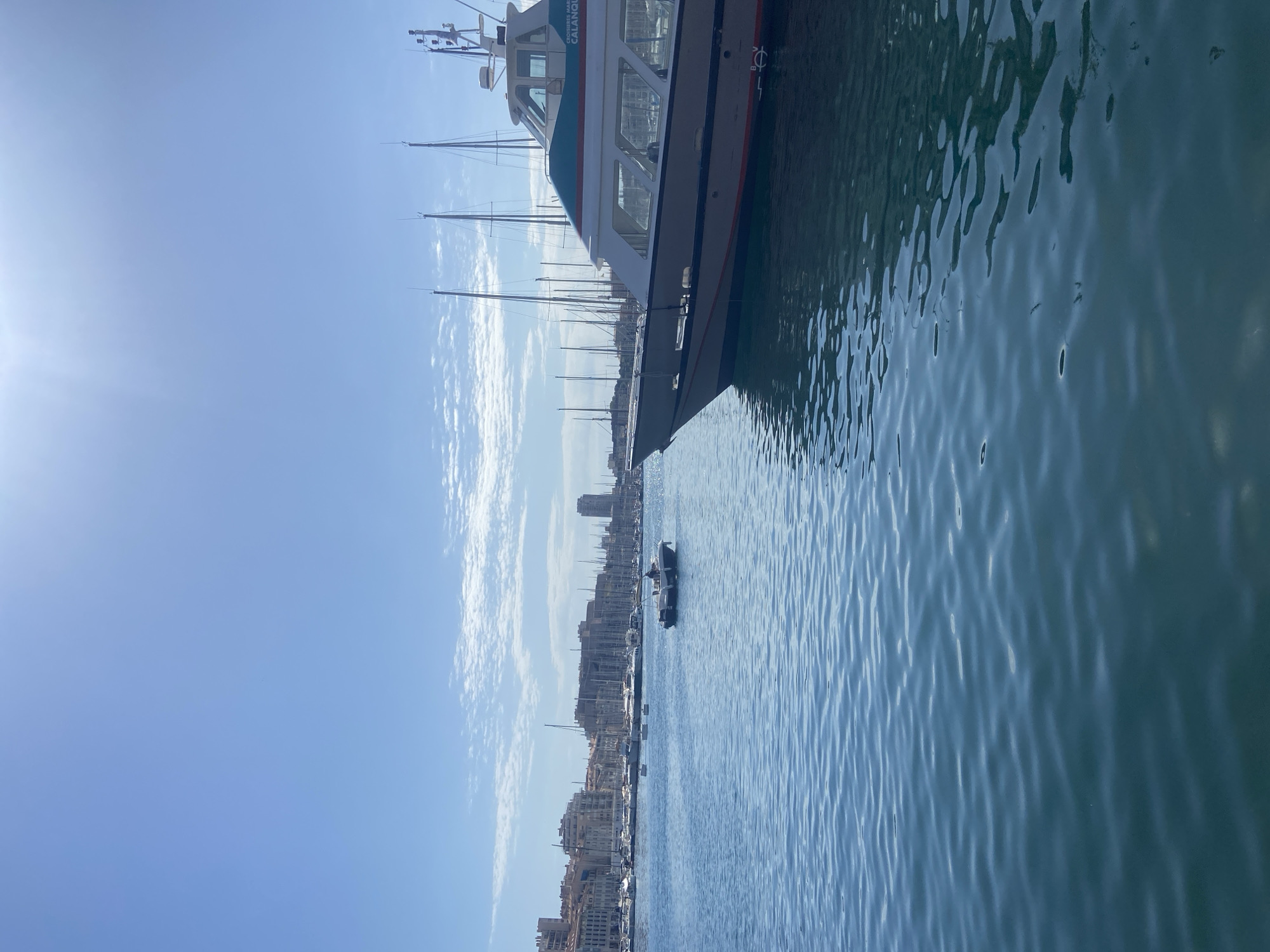 This is a medium shot, eye-level, outdoor, vertical photograph taken in a harbor. The foreground is dominated by calm blue water with subtle ripples. A large white boat with blue and red trim is docked on the right side of the frame, its bow pointing towards the left. The boat has a cabin with windows and the words "CROISIÈRES MARITIMES" and "CALANQUES" are visible on its side. In the middle ground, a small, dark-hulled boat with a single person is moving on the water. Beyond this, a bay is filled with numerous sailboats and other vessels. The background features a city skyline with buildings of varying heights and architectural styles. The sky is a clear, bright blue with a few wispy white clouds and the sun is shining brightly, creating a slight glare in the upper portion of the image. The scene appears to be a sunny day in a coastal city with a busy harbor.