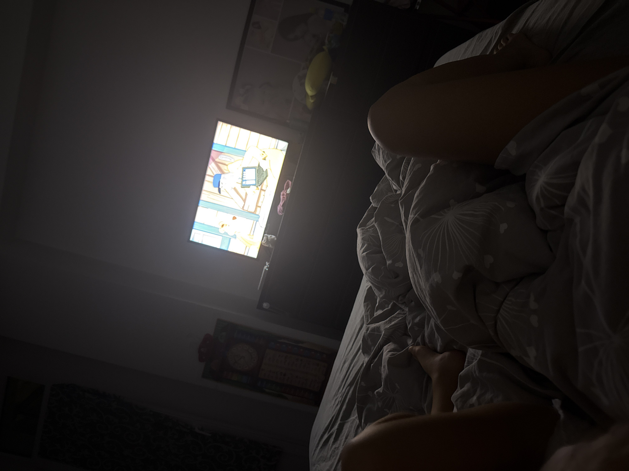In a dimly lit bedroom, a television screen displays a cartoon. The TV is placed on a dark dresser. To the left of the dresser, a grandfather clock stands against the wall. The bed is covered with grey sheets. On the bed, a person's legs are visible, covered by the sheets. The overall impression is one of a quiet evening spent watching television in bed. The image does not provide any specific indicators to place it in Chișinău, Moldova.