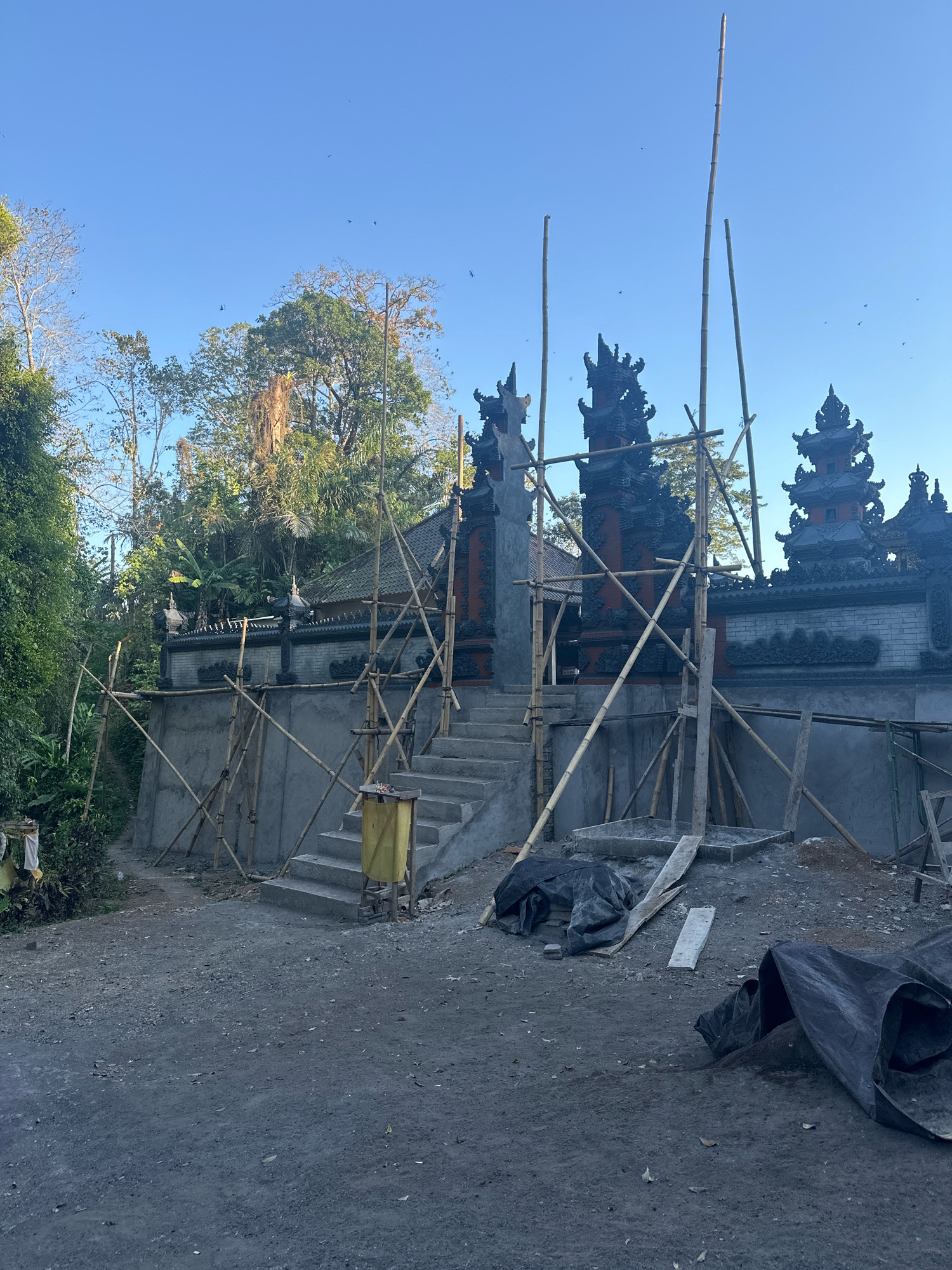 The image captures an outdoor scene of what appears to be a construction site for a traditional Balinese temple gate, possibly in Busungbiu, Indonesia, given the architectural style. The primary structure is a stone gate with intricate dark carvings, flanked by smaller, tiered structures on either side, also adorned with elaborate decorative elements. Bamboo scaffolding is erected around the gate and the surrounding wall, indicating ongoing construction or renovation. Concrete stairs lead up to the central gate. A plain concrete wall extends to the sides of the gate, partially covered by the scaffolding. The ground in front of the gate is a rough, unpaved surface composed of gravel and dirt. Scattered on the ground are black tarps, some covering materials like sand or cement, and pieces of wood. A yellow trash bin with a lid is placed on one of the stairs. The background features lush green foliage and trees, characteristic of a tropical environment, under a clear blue sky. There are no people visible in the scene. The overall impression is one of a developing or restored cultural site.