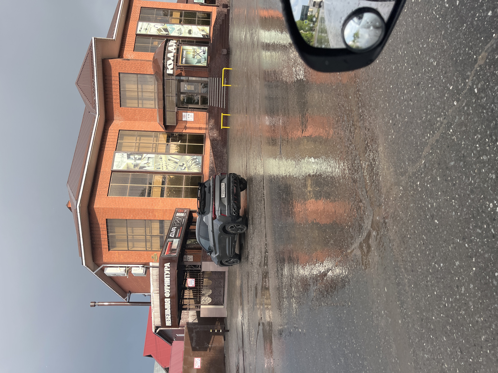 An exterior shot depicts a commercial building and its surroundings, seen from the perspective of a vehicle, with the ground visibly wet from recent rain. The scene is captured in daylight.
The main subject is a multi-story building constructed of reddish-brown brick, featuring a brown roof and numerous large, reflective windows. A prominent entrance is covered by a semi-circular, dark-colored canopy. Above the entrance, a sign in Cyrillic reads "КУЛАЙ" (KULAI), with smaller text below stating "СТУЛЬЯ СТОЛЫ" (Chairs Tables). To the left side of the building, a large vertical sign advertises "МЕБЕЛЬНАЯ ФУРНИТУРА" (Furniture Fittings/Hardware) and includes an "EVA" logo. Small signs near the entrance display information, including business hours, with one showing "9-18".
In front of the building, the ground is a wet asphalt or pavement surface, reflecting the building's facade and the bright, overcast sky. Several yellow bollards or barriers are arranged near the building's entrance. A dark grey SUV is parked on the wet ground, its rear visible, bearing a license plate with the characters "Т 446 КН 98". The '98' region code on the license plate indicates Saint Petersburg, Russia.
The upper right portion of the image shows a black side mirror of the vehicle from which the photograph was taken, with a smaller convex mirror attached, partially reflecting the sky and some foliage. No human activity is visible, and the overall mood is neutral.