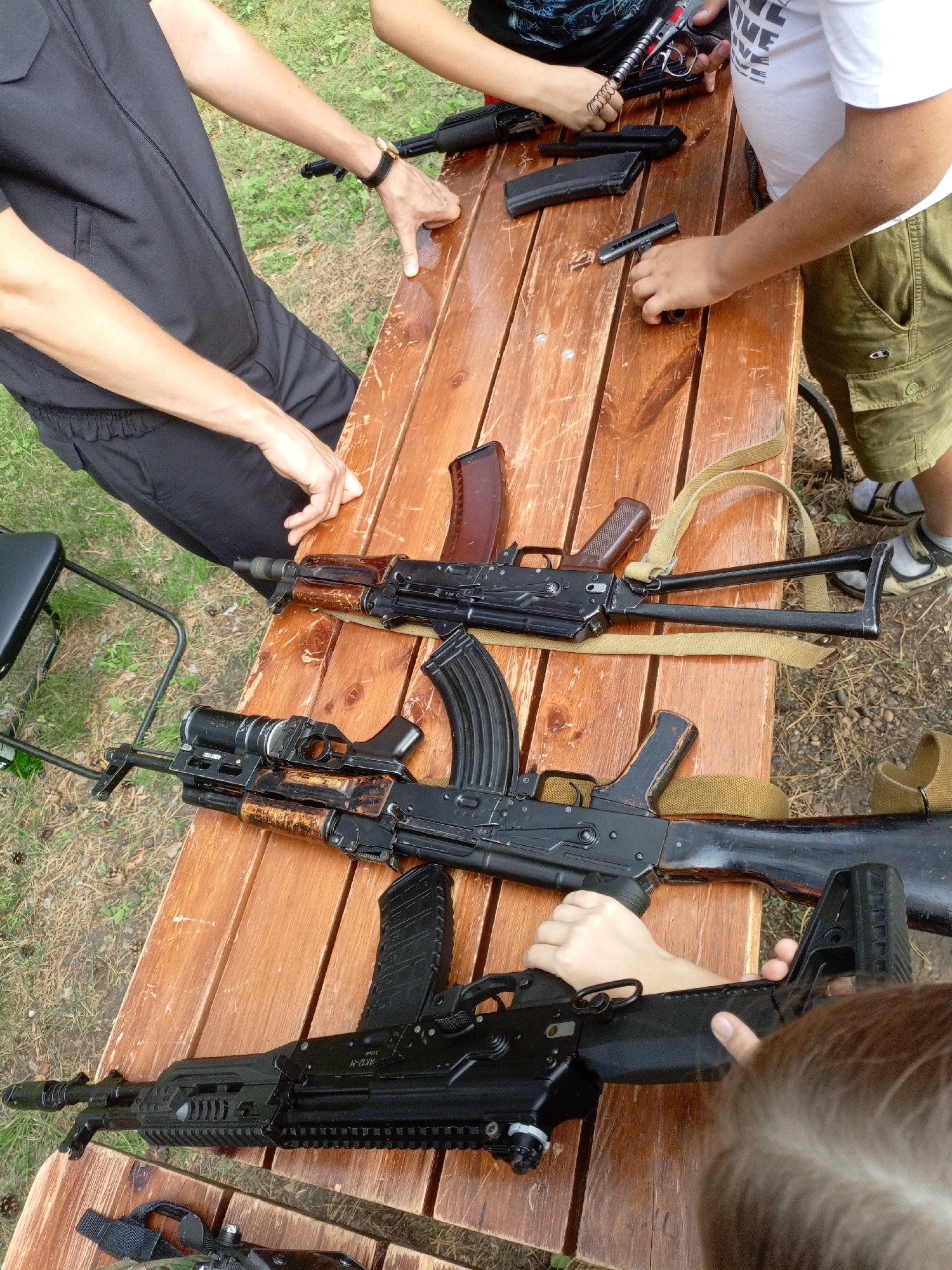 On a wooden table outdoors, several individuals are gathered around firearms, presumably in Zheleznogorsk, Russia. Two individuals, one wearing black and the other a white t-shirt and olive green cargo shorts, are actively handling parts of the weapons. The person in black is wearing a black vest over a black t-shirt and has a watch on their left wrist. The person in the white t-shirt is holding a disassembled firearm component. A third individual, a child, is partially visible in the foreground, their back to the camera, reaching towards a firearm. The table is cluttered with disassembled rifle parts, including magazines, receivers, and barrels. The ground around the table is covered with grass and dirt, with scattered pine cones.