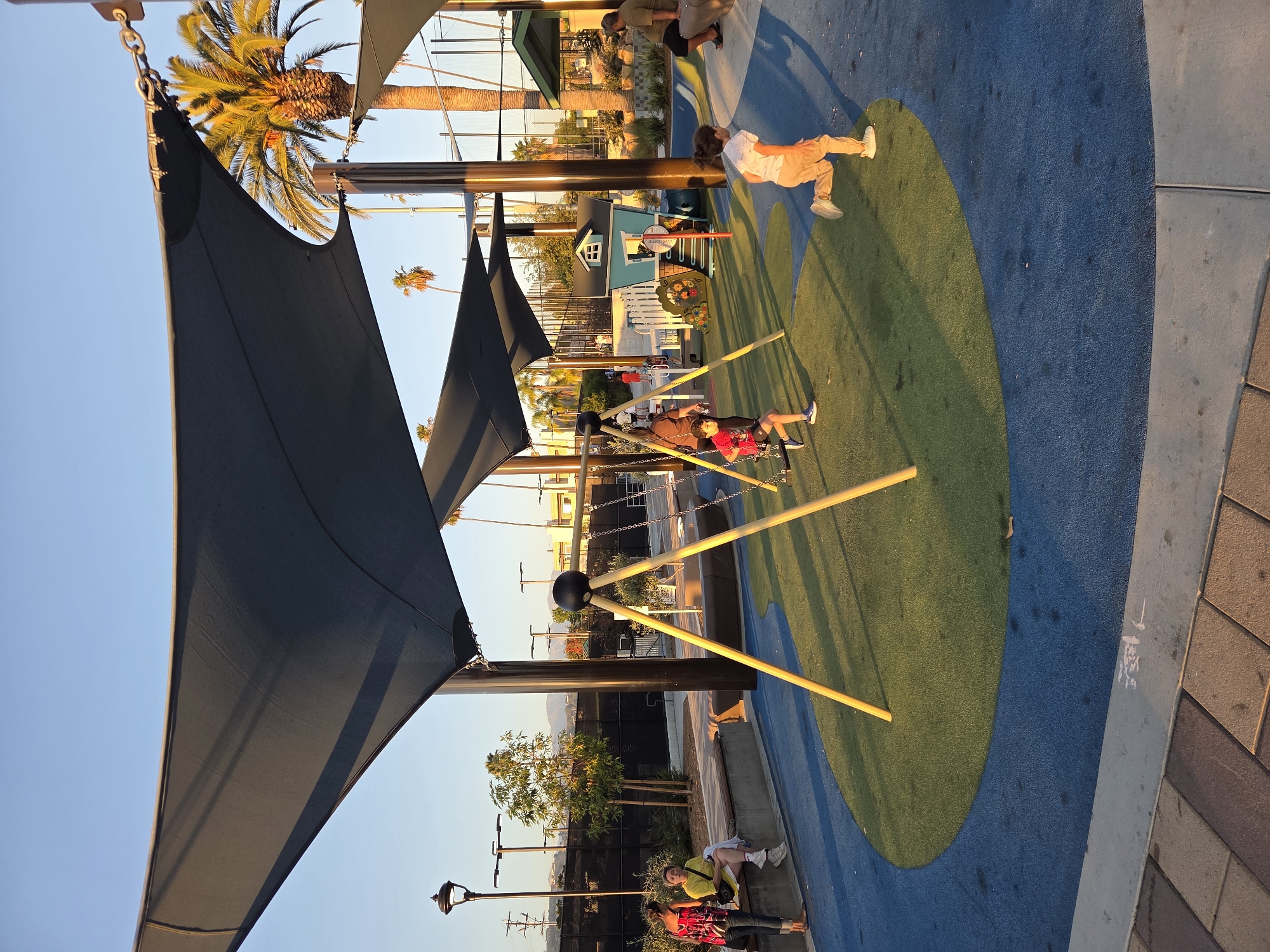In Glendale, United States of America, a playground is depicted with children engaging in various activities under a large, dark blue shade structure. In the foreground, a girl with long dark hair is running from right to left on a blue and green rubberized surface. To her left, a boy with dark hair and wearing a red shirt is on a swing. Behind them, a small teal-colored playhouse with a white picket fence is visible. Two women are seated on a concrete bench to the far left, one wearing a yellow shirt and the other a patterned top. Palm trees and a chain-link fence are visible in the background, along with some buildings and hills. The sun is low in the sky, casting long shadows and illuminating the scene with warm light.