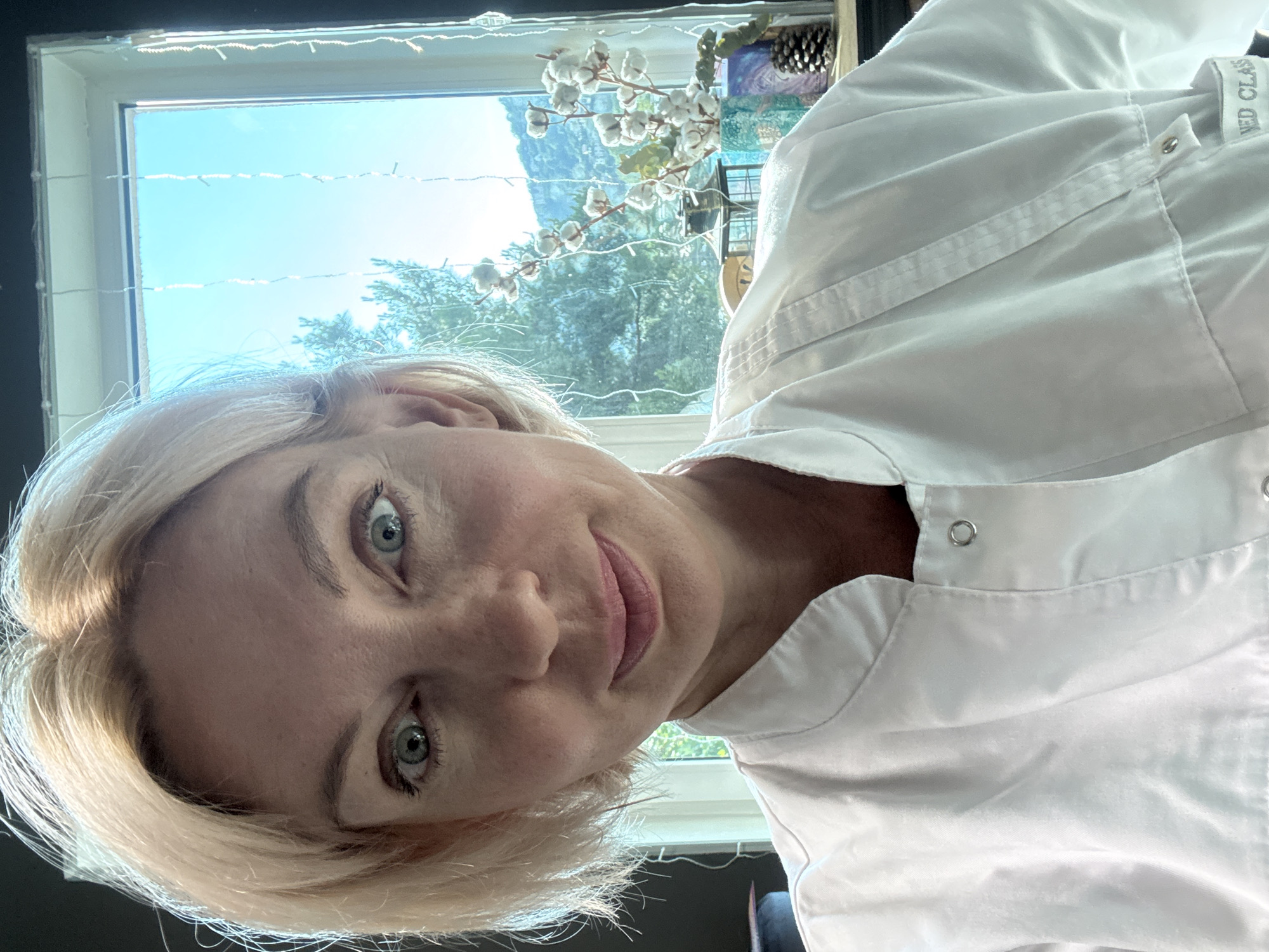 A middle-aged Caucasian woman with blonde hair is pictured in a selfie. She is wearing a white lab coat. The woman is looking directly at the camera with a slight smile. Her eyes are blue. Behind her, a window offers a view of trees and a bright blue sky. Decorative string lights are hanging in front of the window. On a windowsill to the right, there are cotton branches in a vase, a lantern, and a pine cone. The image appears to be taken indoors, likely in a medical or scientific setting, given the lab coat. The presence of greenery and natural light suggests a daytime shot. The background scenery of trees and the bright sky hints at a pleasant climate. No specific location within Sochi, Russia is identifiable from the visual cues in the image.