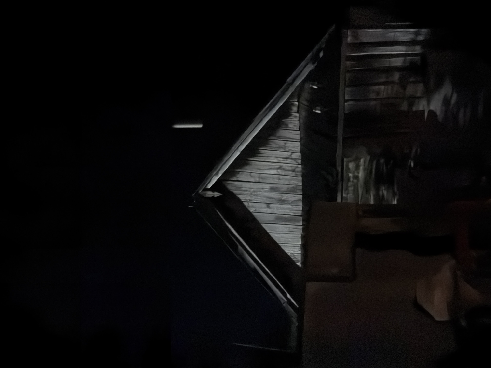 The image displays a nighttime exterior view of a rustic wooden building in Akkuzovo, Russia, under extremely dim illumination. The central and right portions of the frame are dominated by a gabled structure. The gable end features vertically oriented, weathered wooden planks rendered in shades of grey, indicating texture despite the low light. A dark, angled roofline with a discernible peak rises above, its edges faintly catching light. Below the gable, the building's lower section is darker and less defined, with vertical elements visible on the right, possibly indicative of a facade or structural supports. The immediate foreground on the left and center consists of dark, blocky, ill-defined shapes. The upper portion of the image shows an undifferentiated black night sky. A slender, vertical, light-colored object is visible in the upper right against the dark sky, appearing suspended; its specific nature is indeterminate. No individuals or discernible actions are present. The scene is characterized by low light conditions, producing soft contrasts and deep shadows, primarily highlighting the architectural features of the wooden building.