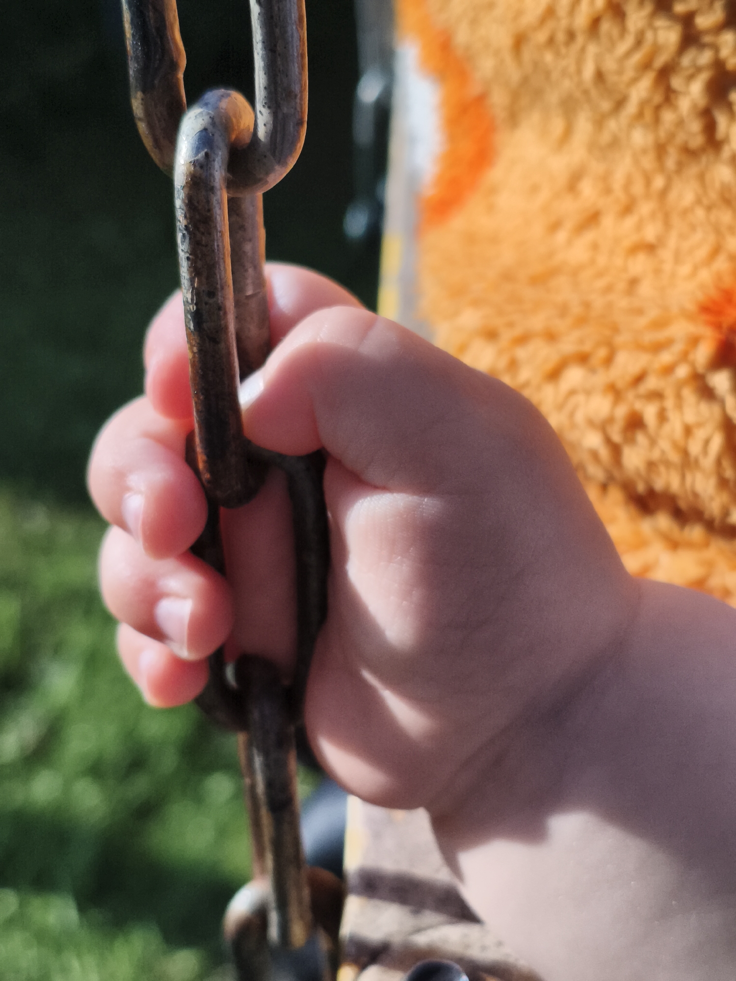 A close-up, angled shot focuses on a small child's hand tightly gripping a rusted metal chain. The chain links are dark brown and worn, with visible signs of age and use. The child's fingers, plump and with small fingernails, are wrapped around one of the links. The skin of the hand appears fair and smooth, with soft light casting shadows across it. In the background, out of focus, is a patch of vibrant green grass, suggesting an outdoor setting, possibly a park or playground. To the right, a fuzzy, bright orange material with darker orange patterns is visible, perhaps a piece of clothing or a toy. The lighting suggests a sunny day, with bright highlights on the chain and the child's skin. This image was captured in Chaikovsky, Russia.