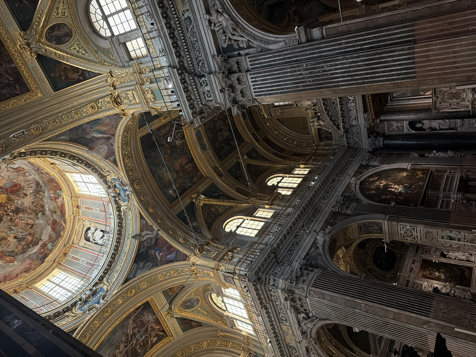 The image shows the interior of a church, likely a cathedral, in Genoa, Italy. The view is angled upwards, showcasing the intricate ceiling. The architecture is ornate, featuring gold leaf accents, detailed paintings, and classical motifs. Windows are integrated into the design, allowing natural light to illuminate the space. There are no visible people in the image. The overall impression is one of grandeur and historical significance.