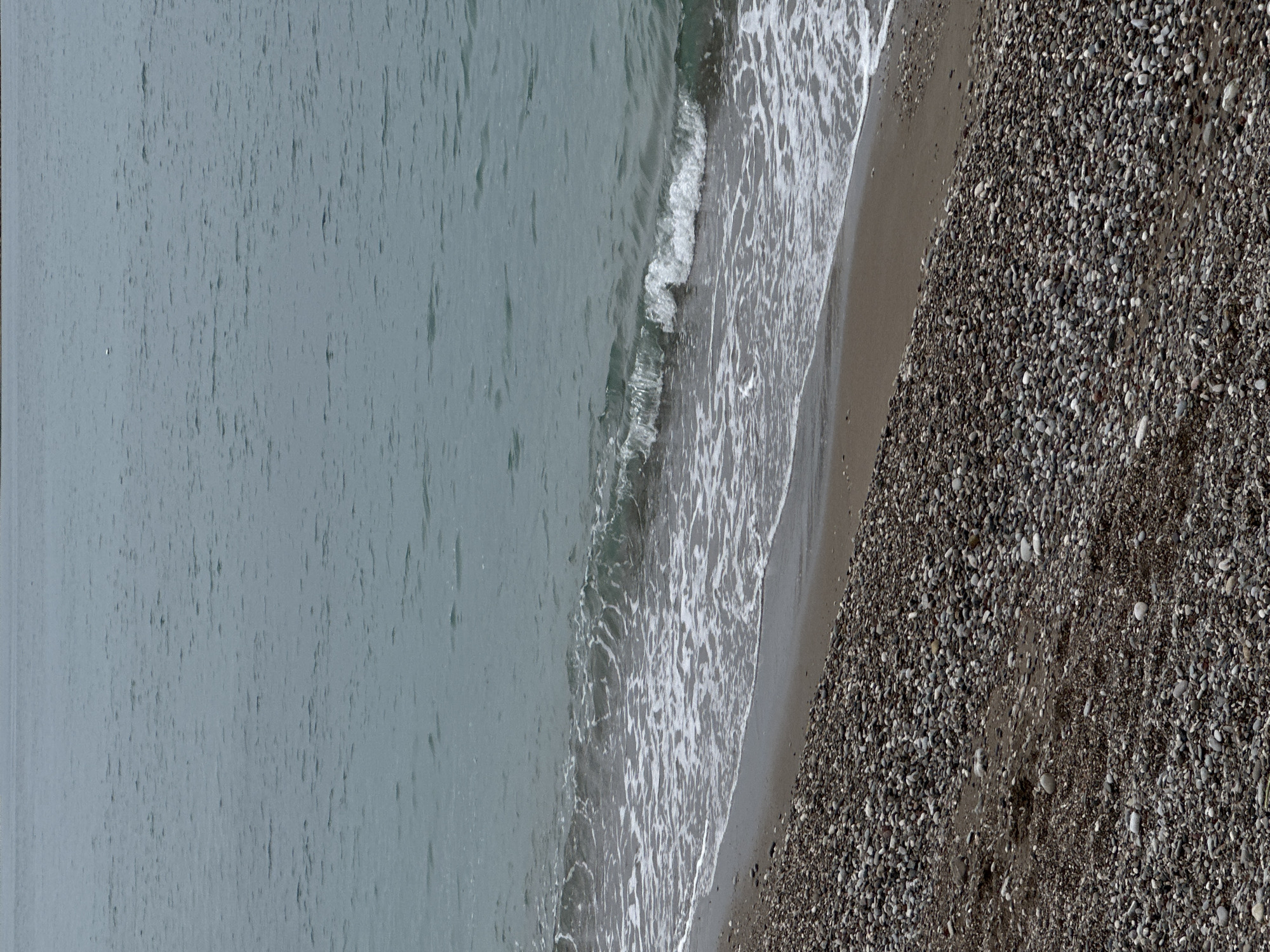 The image depicts a view of the coastline in Bar, Montenegro. The scene consists of three primary elements: the sea, the shoreline, and the pebble beach. The water is a grayish color and appears calm with some slight ripples and small waves. The shoreline has white, foamy waves meeting the dark pebble beach. The beach is densely covered with pebbles.