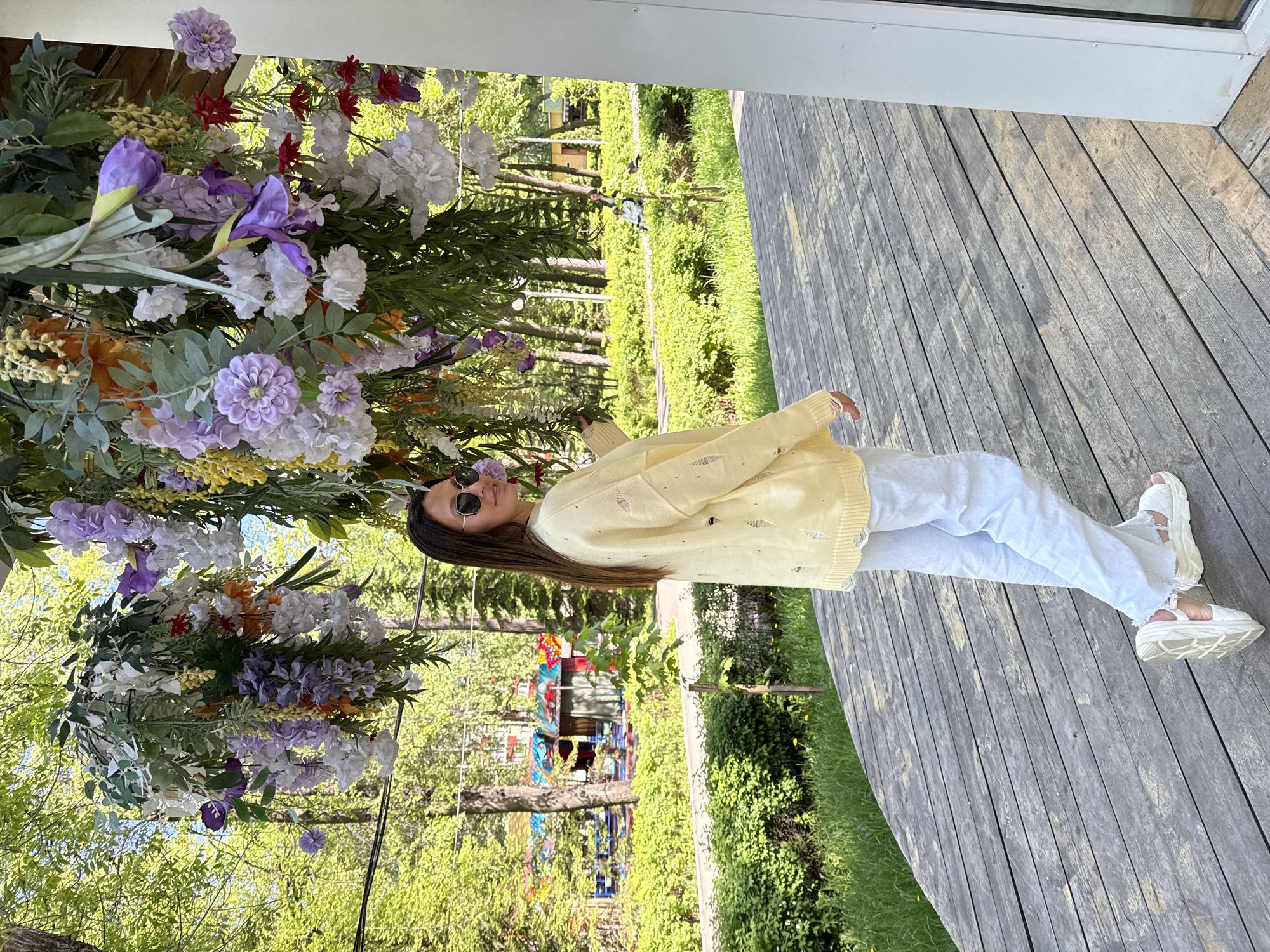 A woman with long, dark hair and sunglasses stands on a wooden deck, partially framed by a vibrant floral arrangement featuring purple, white, and orange flowers. She wears a light yellow sweater and light-colored pants, paired with white sandals. In the background, a green, park-like environment is visible, including trees and a colorful structure resembling a playground or art installation. This photograph was taken in the city of Perm, Russia.