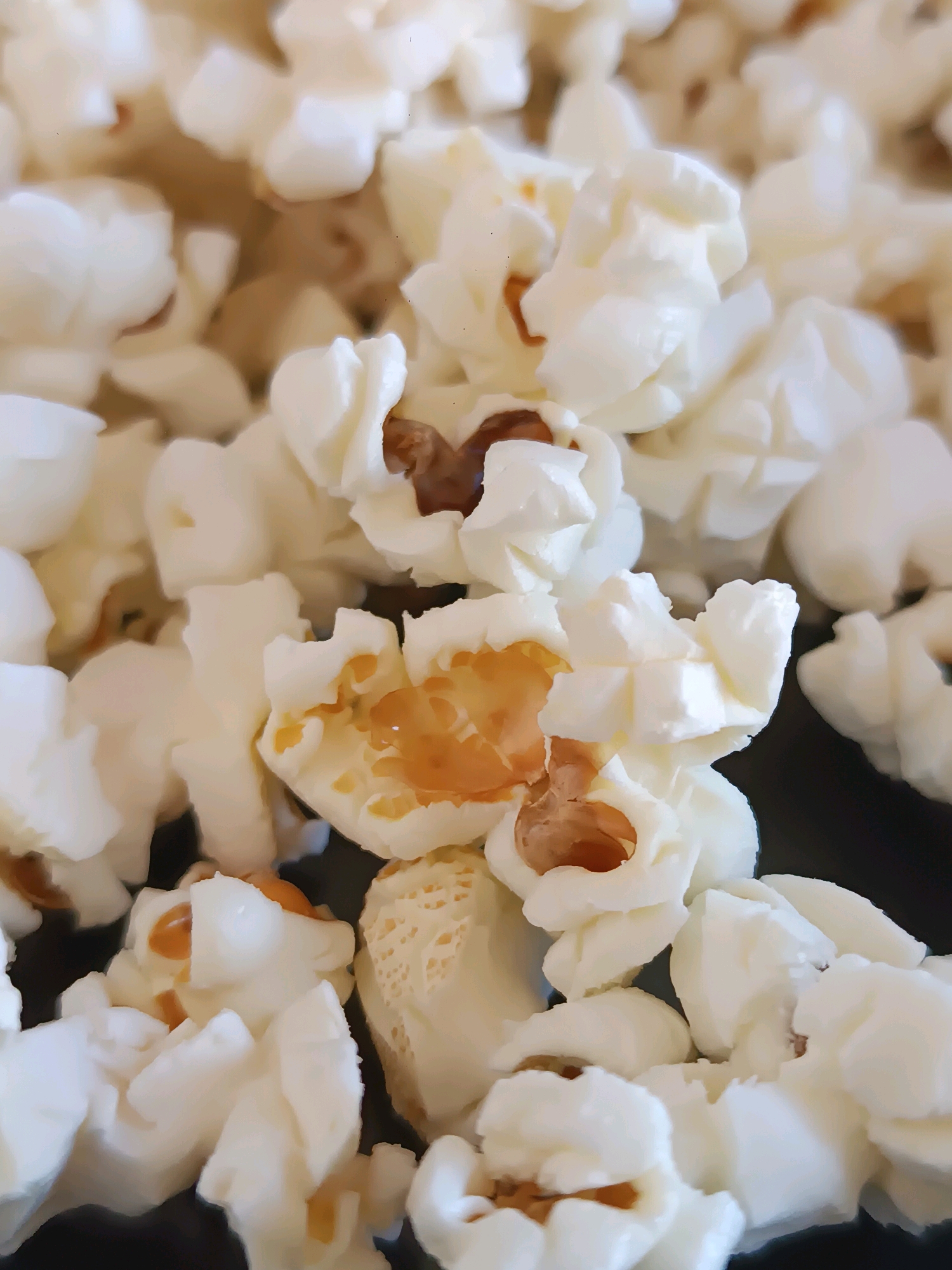 The image is a close-up, overhead shot of a pile of freshly popped popcorn. The popcorn is predominantly white with some kernels showing golden-brown hues in the center, indicating they have been popped. The texture appears fluffy and irregular, characteristic of popcorn. The popcorn is piled on a dark surface, likely a bowl or plate, which is only partially visible at the bottom and right edges of the frame. The lighting is bright, highlighting the texture and shapes of the popcorn. There are no people or discernable environmental features beyond the popcorn and the dark surface. The scene is devoid of any specific context suggesting a location within the city of Viadana, Italy.