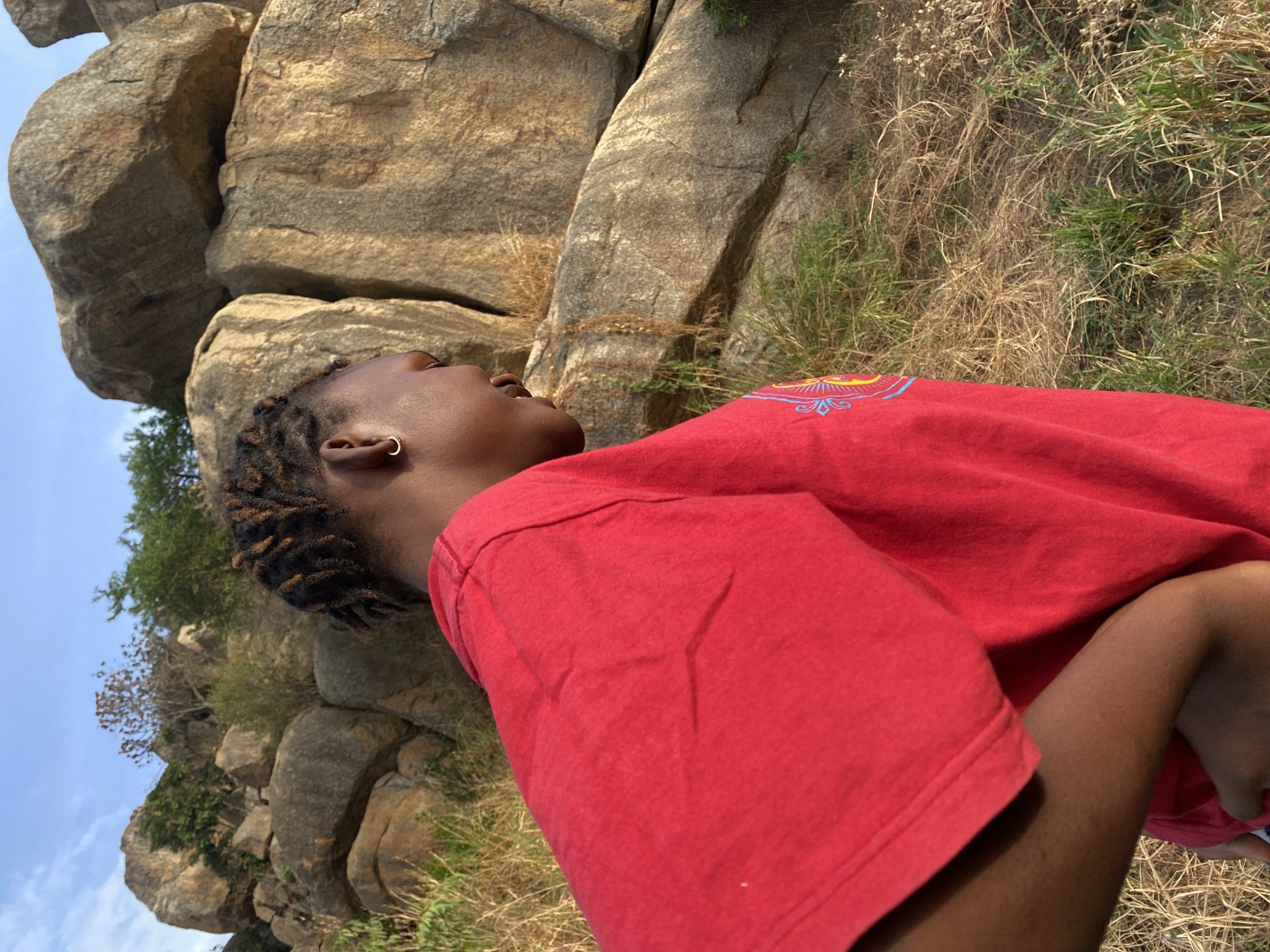 A person wearing a red t-shirt with a colorful design is outdoors. They are positioned in front of large, light-colored rocks and some green and brown vegetation. The sky is visible and clear. The location is near Vom, Nigeria, as Vom is known for its rock formations. The person's face is tilted upwards. They are wearing an earring.