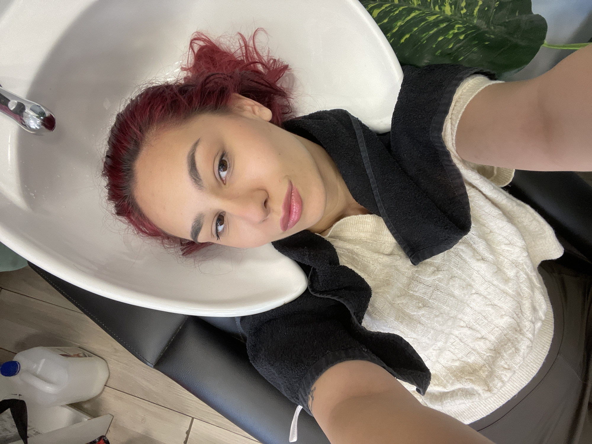 A young woman with vibrant reddish-pink hair is captured in a selfie-style photograph while lying in a white salon shampoo bowl. Her head is tilted back into the basin, suggesting she is having her hair washed or treated. Her roots show a darker, natural hair color. She looks towards the camera with a relaxed expression and a faint smile, wearing natural-looking makeup with defined eyebrows and a pink lip color.
A black towel is draped around her neck and shoulders, providing protection from water. She is wearing a light-colored, possibly off-white, cable-knit sweater. The white ceramic of the shampoo bowl and a chrome faucet are prominent in the foreground. Her body rests on a dark, possibly black, salon chair.
In the background, hints of green plant leaves are visible, suggesting either real foliage or artificial decoration. The flooring beneath the chair appears to be light-colored wood or laminate. A white plastic bottle with a blue cap is visible on the floor next to the chair. The setting appears to be an indoor hair salon or a similar professional beauty environment. The lighting suggests it is daytime. No discernible text is visible in the image.