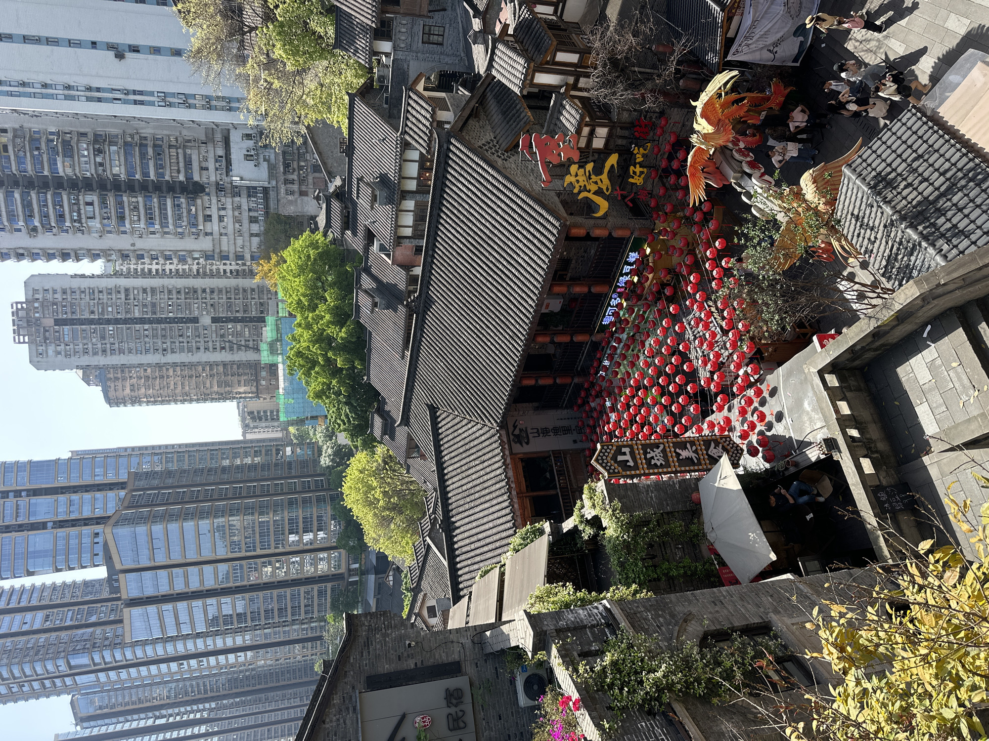 The image, taken from an elevated perspective, features a scene in the Yuzhong District of Chongqing, China. The location is marked by a blend of traditional and modern architecture. Several high-rise buildings with glass windows stand adjacent to traditional Chinese-style buildings with dark-tiled roofs. Strings of red lanterns are strung along the street, adding a festive atmosphere. A large golden bird statue, likely a phoenix, is also present.
The scene includes several people walking or standing around the area, particularly in the open space. Other elements include signage with Chinese characters, trees, and vegetation interspersed among the buildings.
