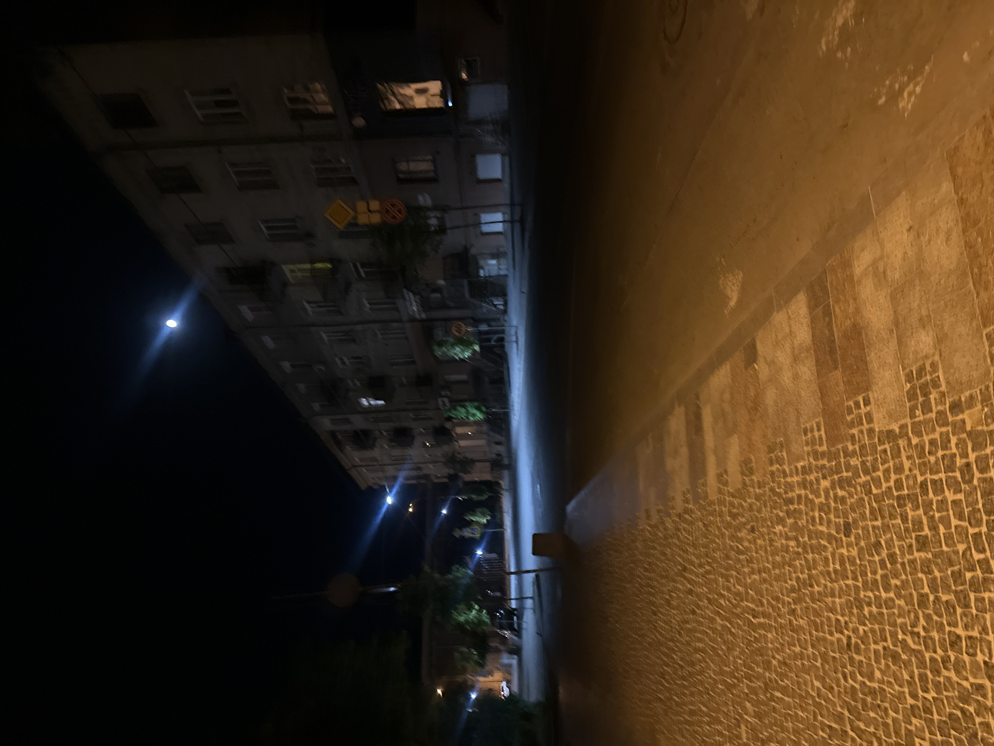 At night, a tall apartment building with numerous windows stands alongside a street in Ternopil, Ukraine. A bright light, possibly the moon, illuminates the top left of the building. Streetlights and building lights are visible. The foreground shows a cobblestone sidewalk, possibly wet, next to a smooth paved area. There are no visible people or distinct actions.