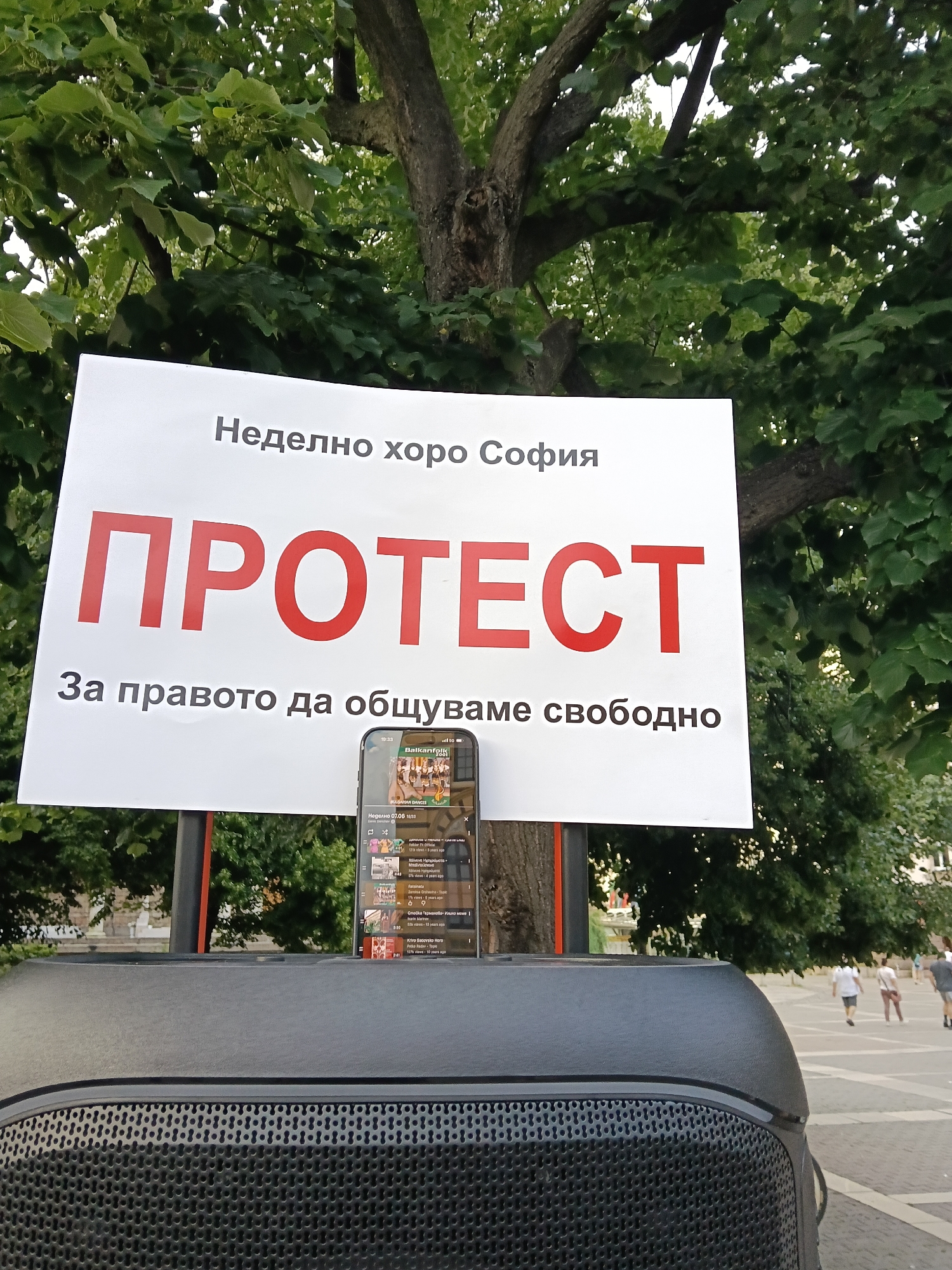 A low-angle outdoor photograph captures a white protest sign, a speaker, and a smartphone against a backdrop of dense green foliage and a public square in Sofia, Bulgaria.
In the foreground, a dark grey speaker with a textured grille is partially visible, occupying the bottom portion of the frame. Resting on the speaker's top surface, a smartphone displays a YouTube interface. The phone screen shows a playlist under the heading "BULGARIAN DANCES," with titles such as "Неделно 07.06" (Sunday 07.06) and various Bulgarian folk dance names (e.g., "Ganka," "Adamo Horo Sofia," "Radomira," "Smolitsa Germanova," "Krivo Sadovsko Horo").
Behind the speaker and phone, a rectangular white sign is mounted on a stand. The sign features text in Bulgarian:
* The top line reads "Неделно хоро София" (Sunday Horo Sofia) in grey capital letters.
* Below it, the word "ПРОТЕСТ" (PROTEST) is prominently displayed in large, bold red capital letters.
* The bottom line states "За правото да общуваме свободно" (For the right to communicate freely) in grey capital letters.
The background consists of lush green deciduous trees with numerous leaves and branches, indicating a park-like or urban green space. A thick tree trunk is visible behind the right side of the sign. In the far background, beyond the trees, a paved public square is visible with several blurred human figures walking. Distant buildings are subtly discernible on the horizon. The lighting suggests daytime conditions.