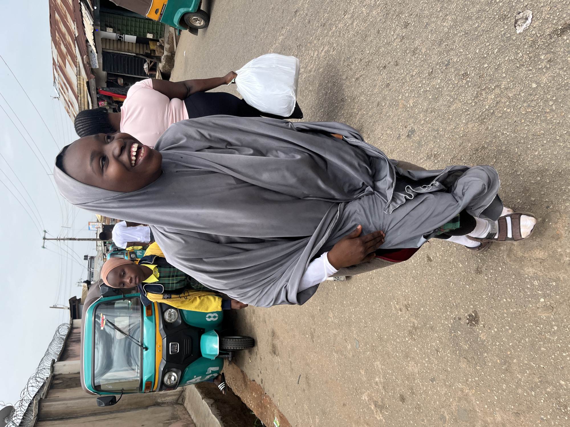 The scene depicts a street in Gura Top, Nigeria. A woman wearing a gray hijab and matching pants is seen smiling at the camera. She is seated on the back of a vehicle (likely a motorcycle or scooter) with brown and white sandals. Another woman in a pink top is walking behind her, carrying a white plastic bag. A light blue and green three-wheeled vehicle ("keke napep" or auto-rickshaw) is parked on the side of the road with a girl wearing a yellow and green jacket riding in the passenger seat. A building with a rusty corrugated metal roof and barred windows is visible in the background. Another auto-rickshaw is partially visible behind the first one. A concrete wall topped with barbed wire runs along the side of the road. The street surface appears to be paved or made of concrete. It is daytime, and the sky is overcast.